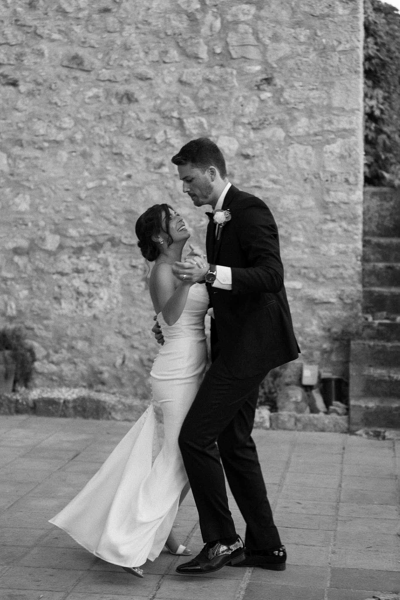 Black and white candid of bride and groom dancing on stone terrace against rustic wall