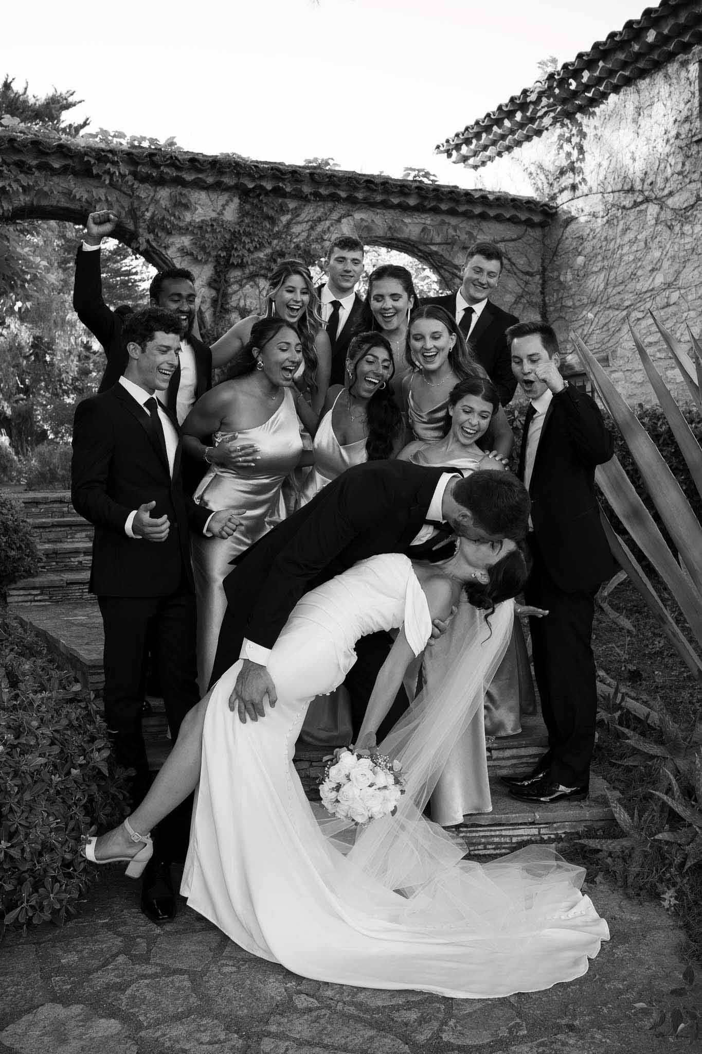 Black-and-white portrait of groom dipping bride while bridal party cheers on stone steps at a Provencal venue courtyard