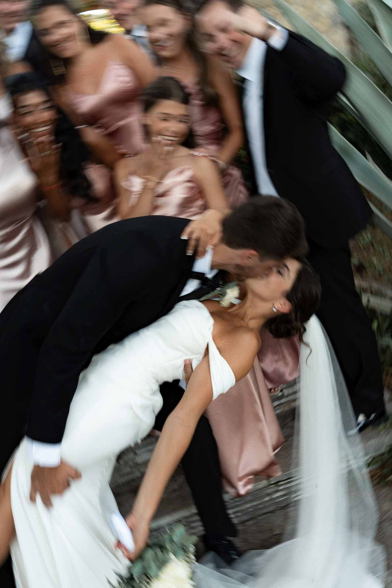 Groom dips and kisses bride surrounded by cheering bridal party in blush dresses and black suits, motion blur effect