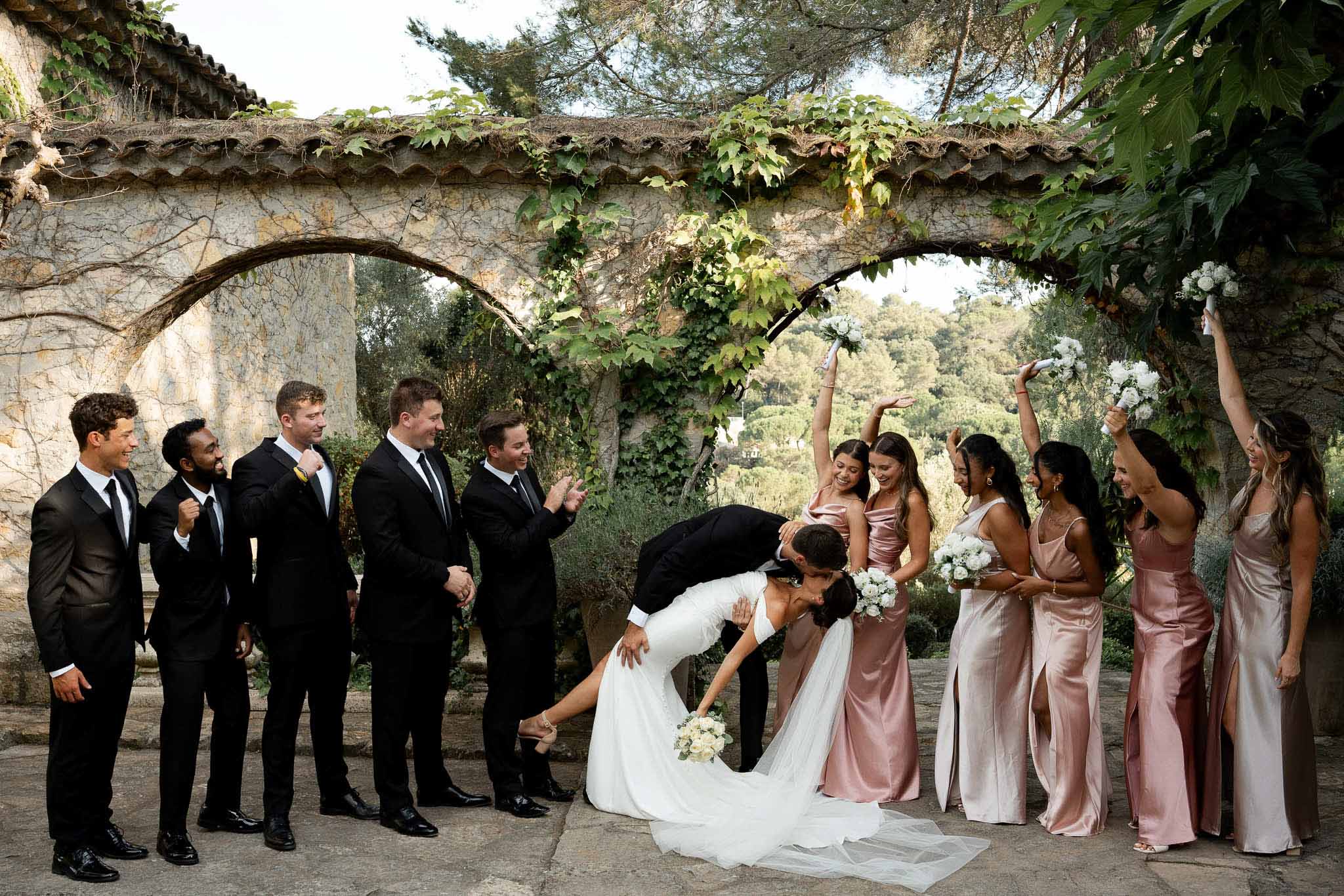 Groom dipping bride as five bridesmaids in blush satin and five groomsmen celebrate under vine archway