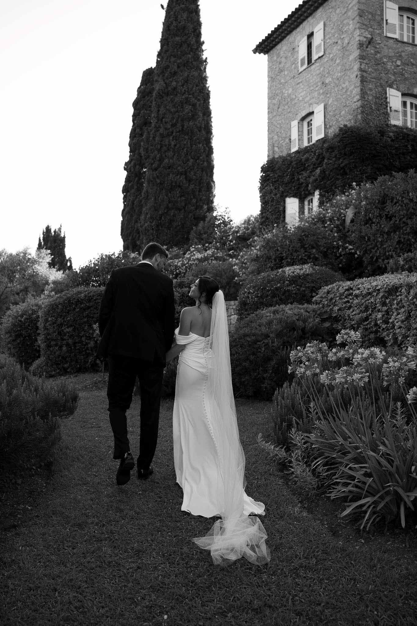 Couple walks away on garden path as bride's cathedral veil trails behind at ivy-clad bastide in B&W
