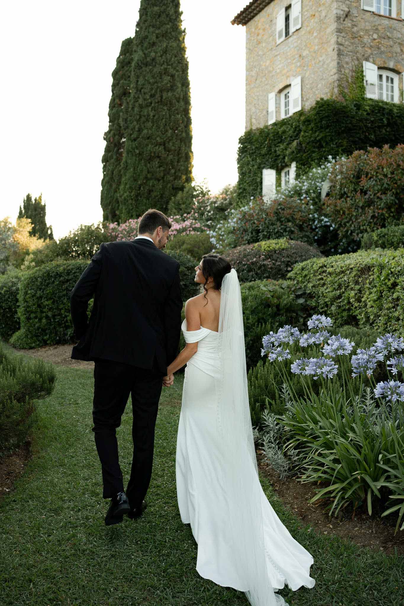 Bride and groom walking hand in hand toward ivy-covered chateau with cathedral veil trailing