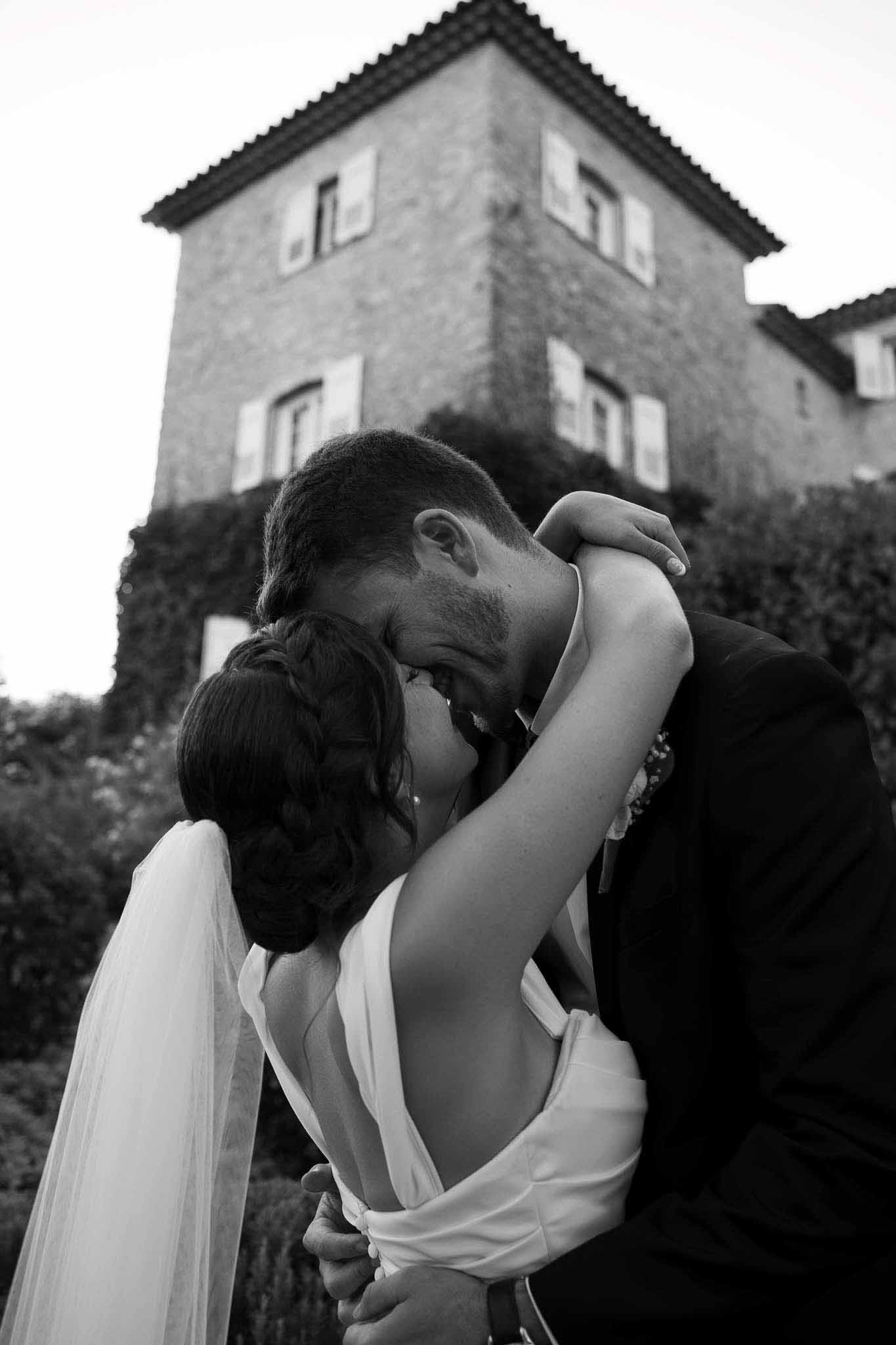 Black and white low-angle couple kissing with flowing veil beside ivy-covered chateau tower with shuttered windows