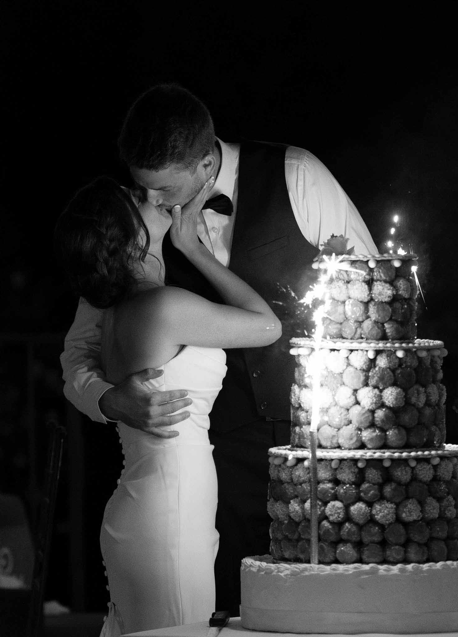 Black-and-white photo of couple kissing beside three-tier croquembouche topped with lit sparklers