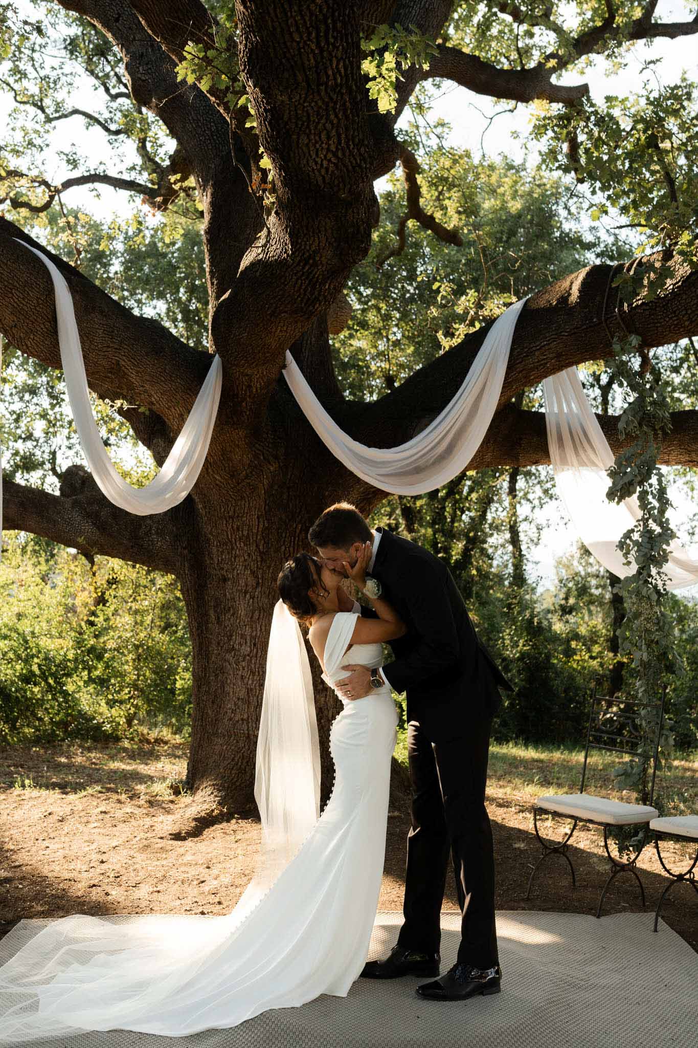 First kiss beneath ancient oak tree draped with white fabric during outdoor garden ceremony