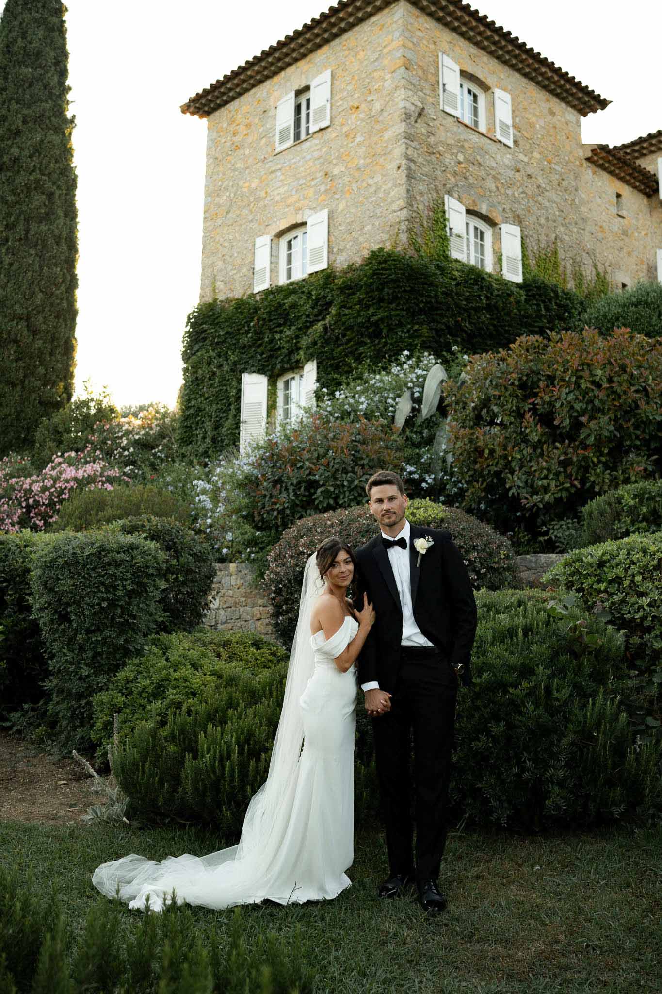 Bride in off-shoulder white gown with cathedral train and groom in black tuxedo holding hands in Provencal garden