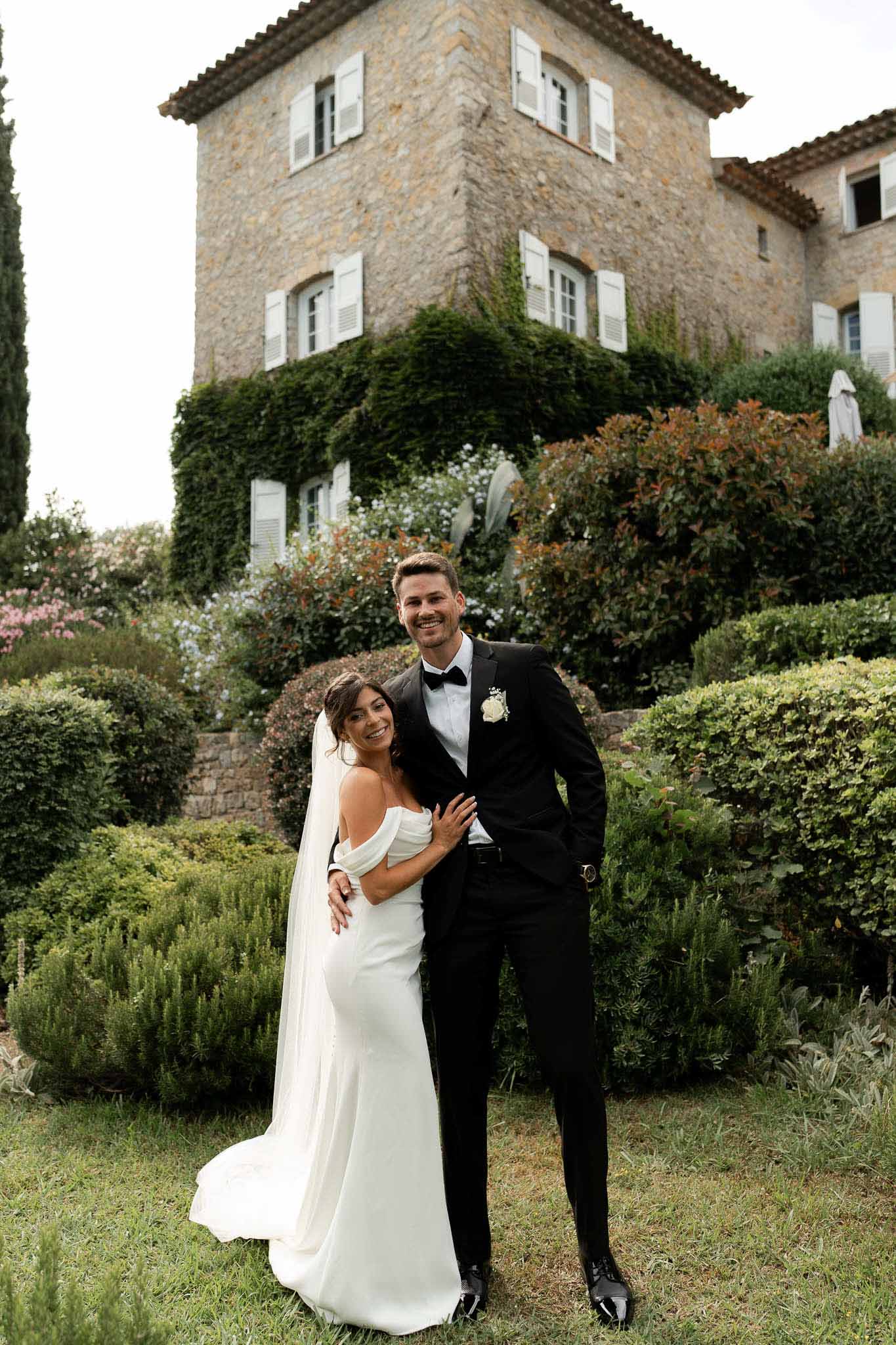 Couple smiling at camera on tiered garden before Provencal stone manor, bride in off-shoulder crepe gown with cape train