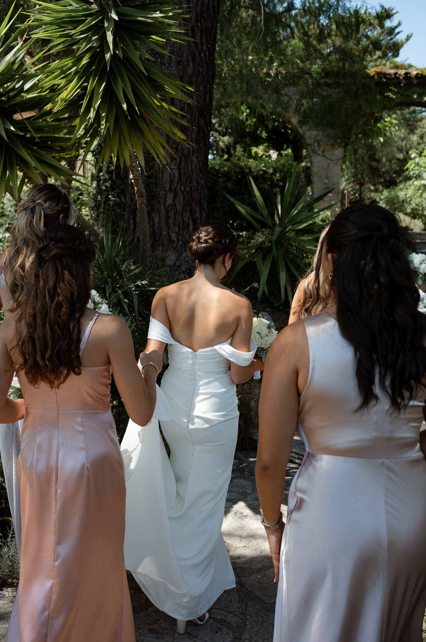 Bride walking from behind with two bridesmaids in blush satin holding train at Mediterranean venue