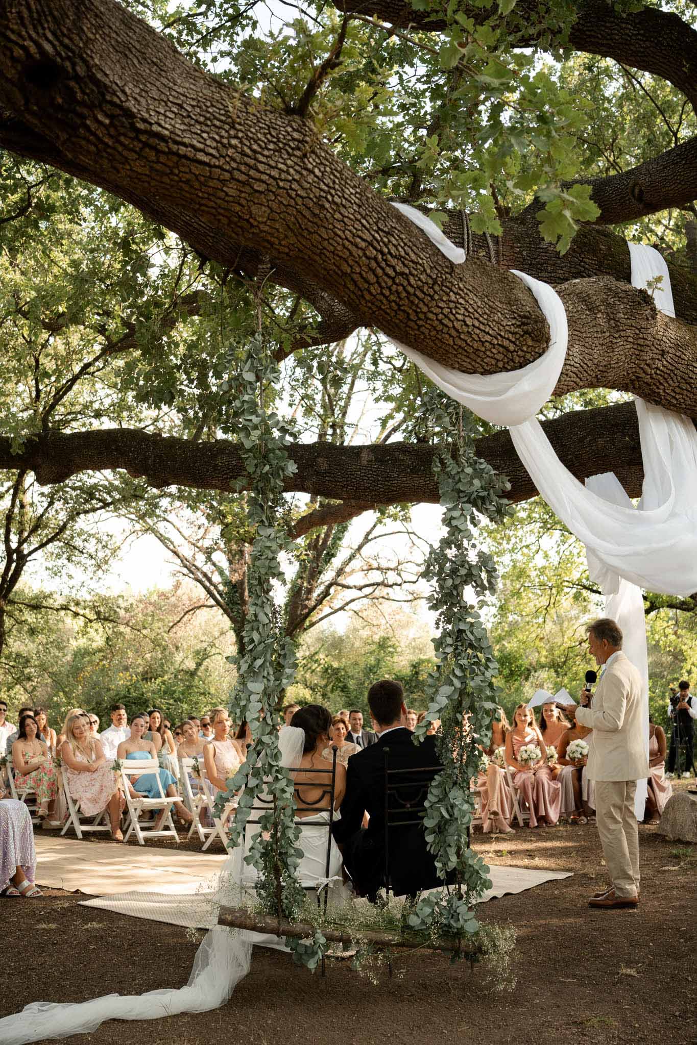 Outdoor wedding ceremony under oak tree with eucalyptus garlands, guests seated in semicircle around couple