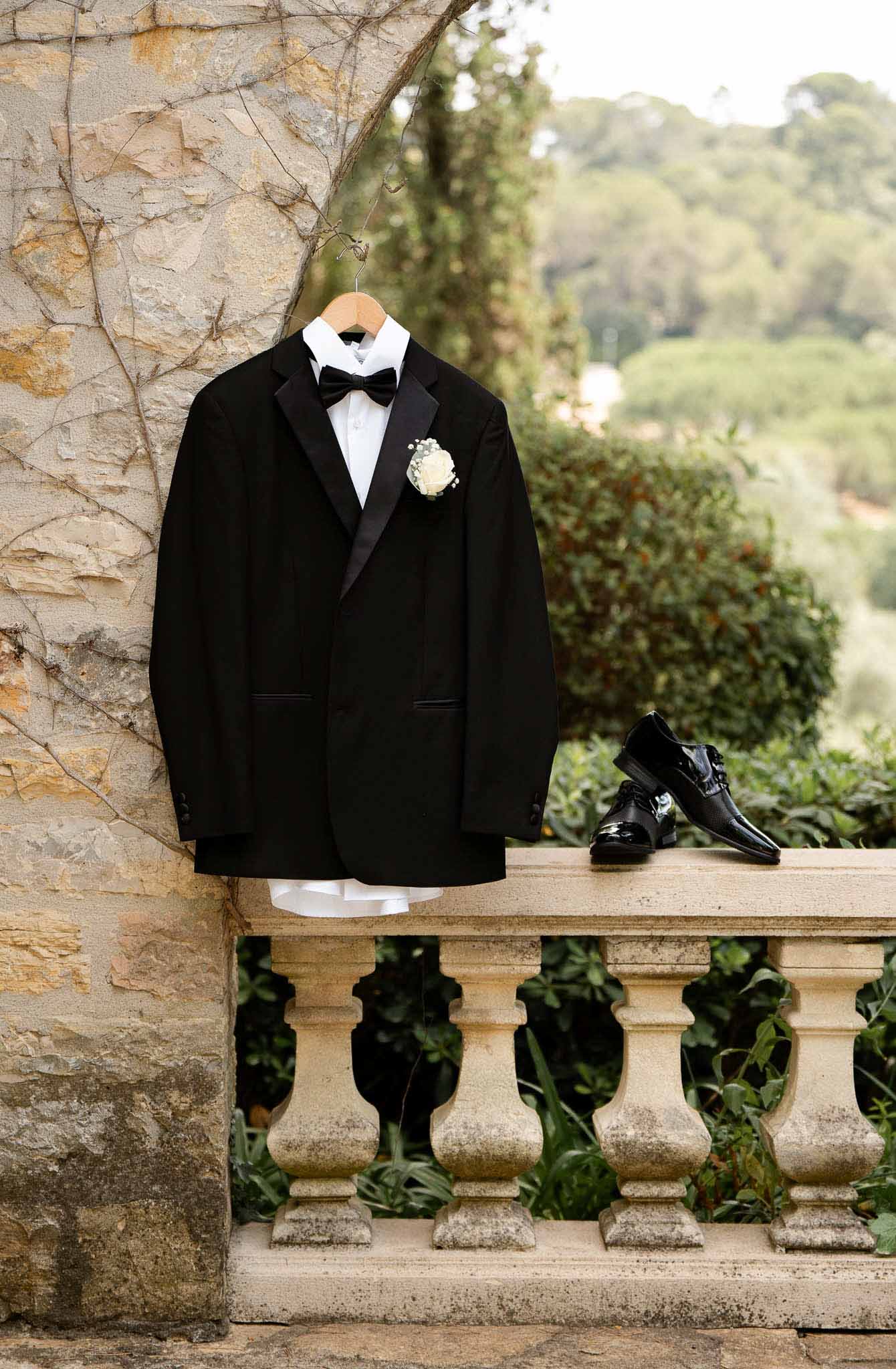 Black tuxedo with satin lapels and ivory rose boutonniere hanging on stone balcony with patent leather shoes