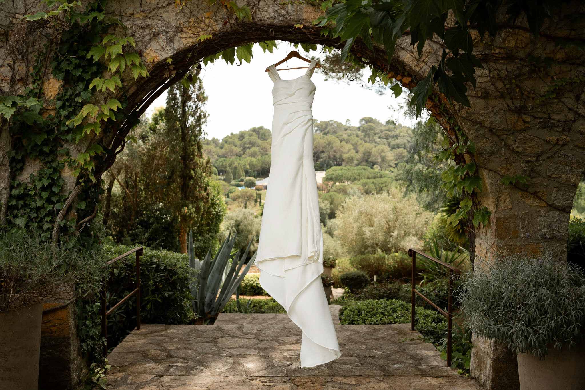 Ivory square-neck gown hanging in stone archway with climbing ivy framing terraced garden view