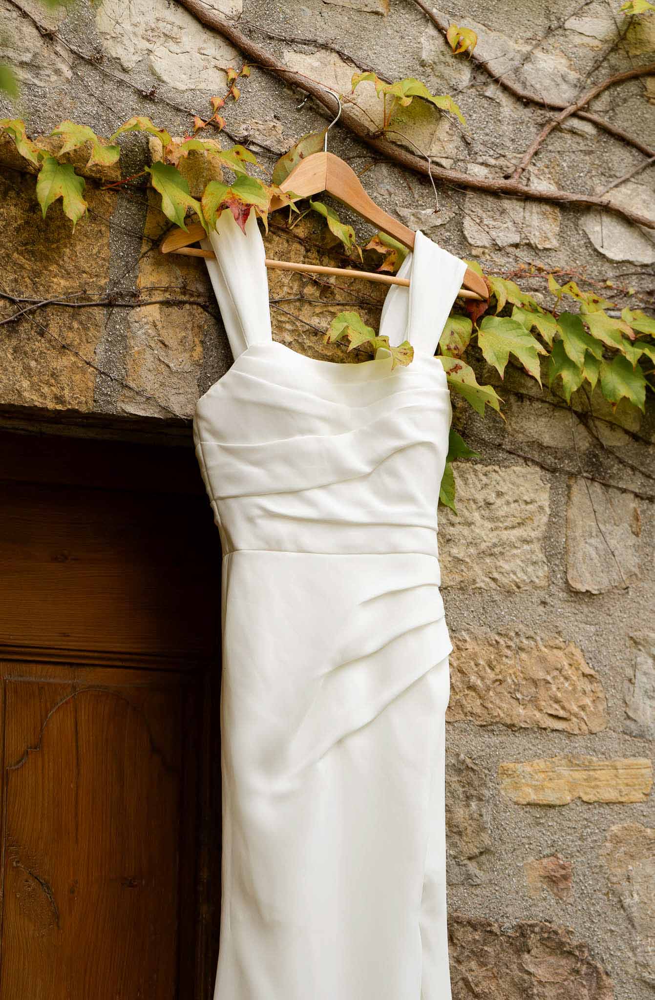 Ivory ruched column wedding dress hanging on wooden hanger against stone building exterior