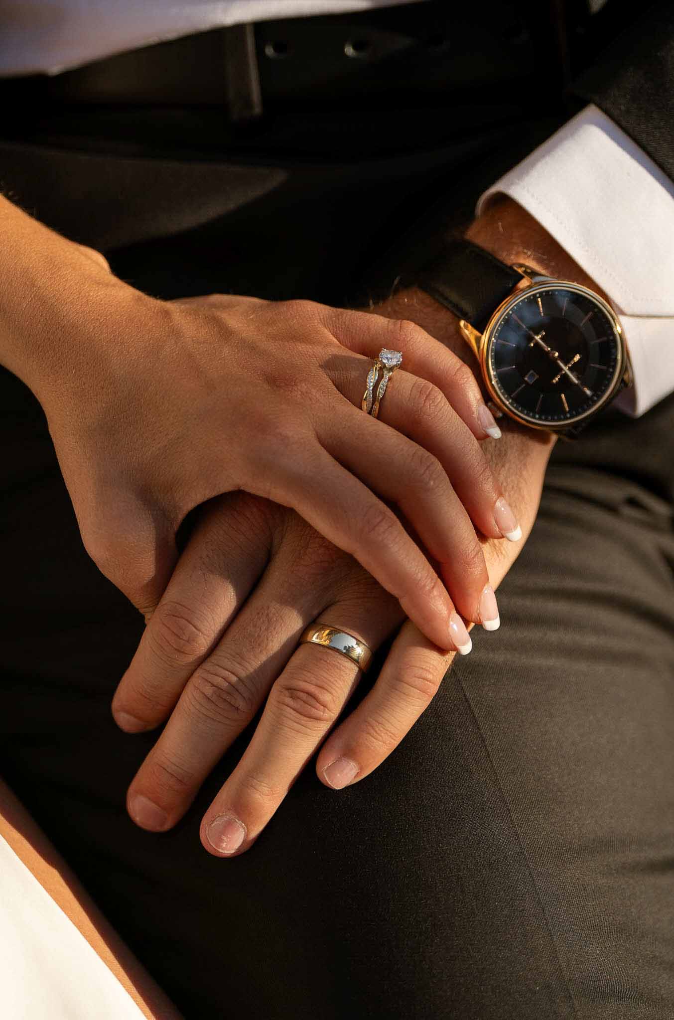 Close-up of bride and groom's hands showing diamond engagement ring, pave wedding band, and gold wedding band with watch