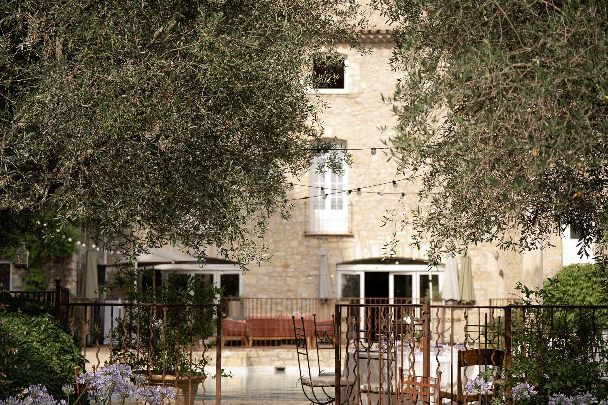 Provencal stone farmhouse with Edison lights and reception tables viewed through olive trees and iron panels