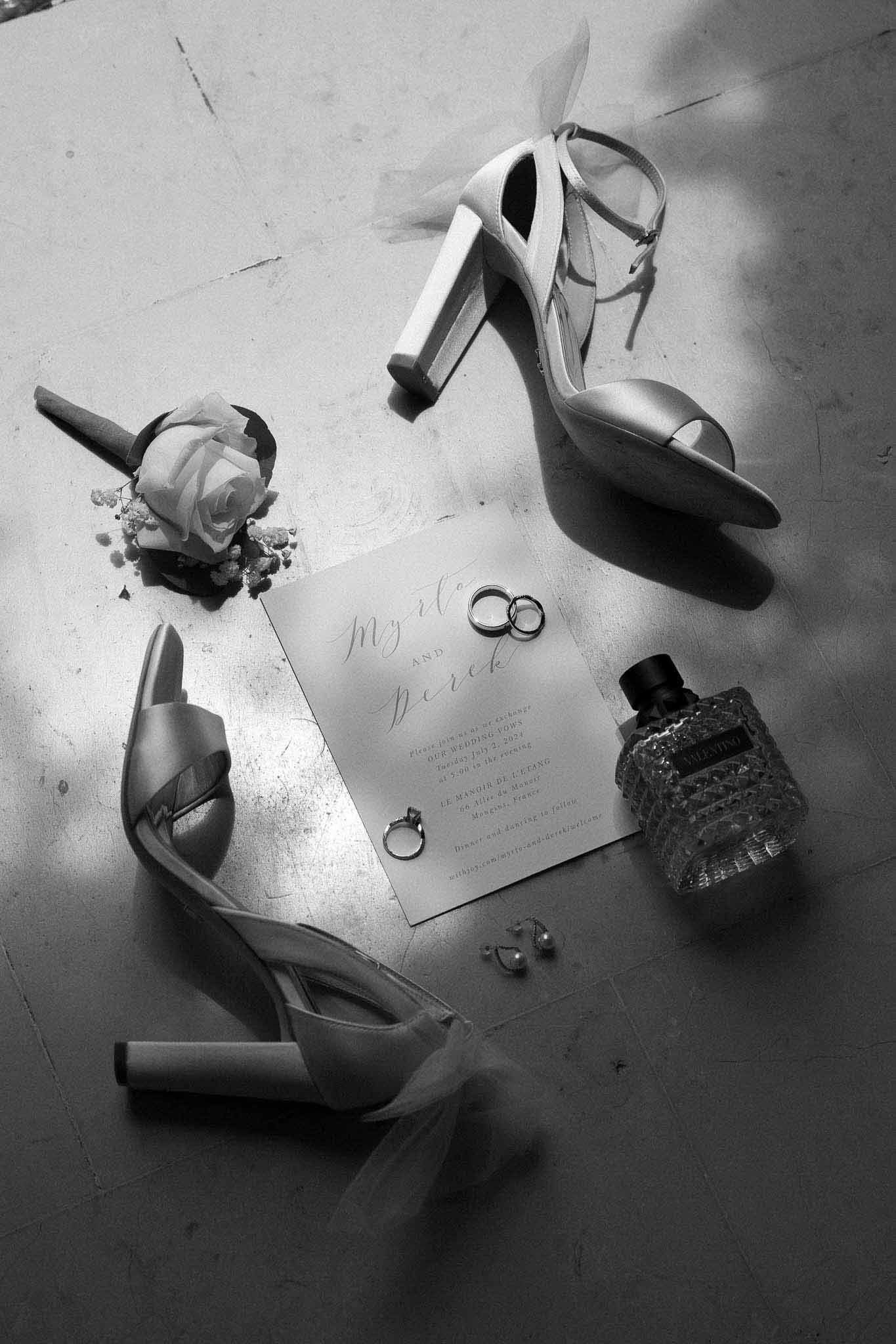 Black-and-white flat lay of bridal shoes, wedding invitation, rings, boutonniere, and Valentino perfume