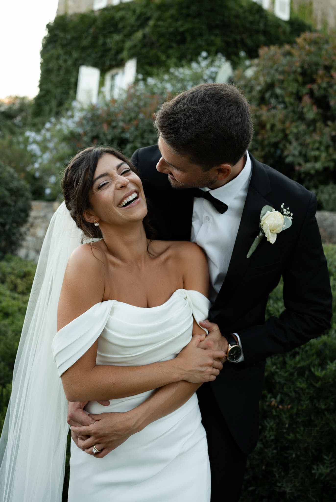 Groom embracing laughing bride in off-shoulder draped gown and pearl earrings before ivy-covered chateau