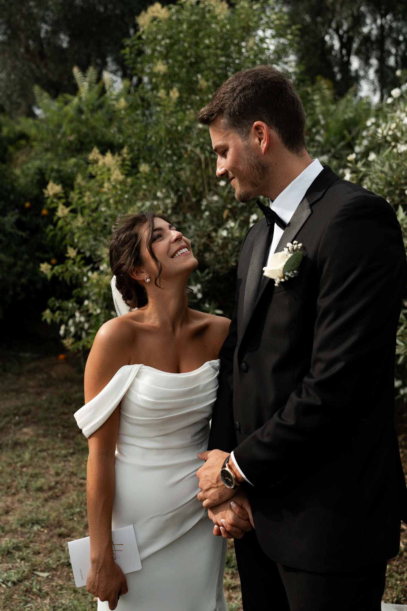 Couple holding hands in garden with bride in cowl-neck satin gown and groom in satin-lapel tuxedo