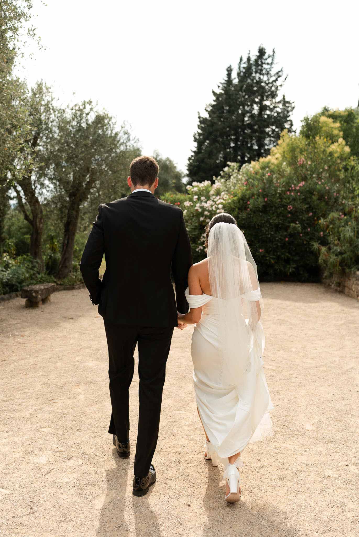 Bride and groom walking hand-in-hand along a garden gravel path seen from behind