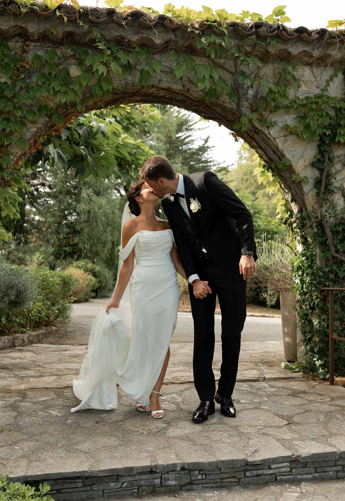 Bride and groom kissing beneath ivy-covered stone archway on paved terrace in garden setting
