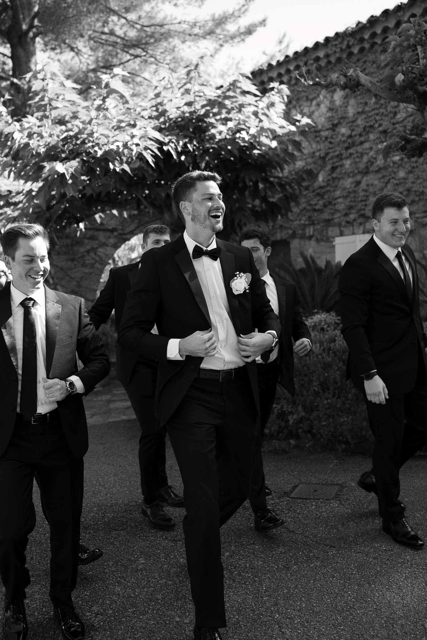 Black and white groom and four groomsmen laughing while walking on path at French stone property