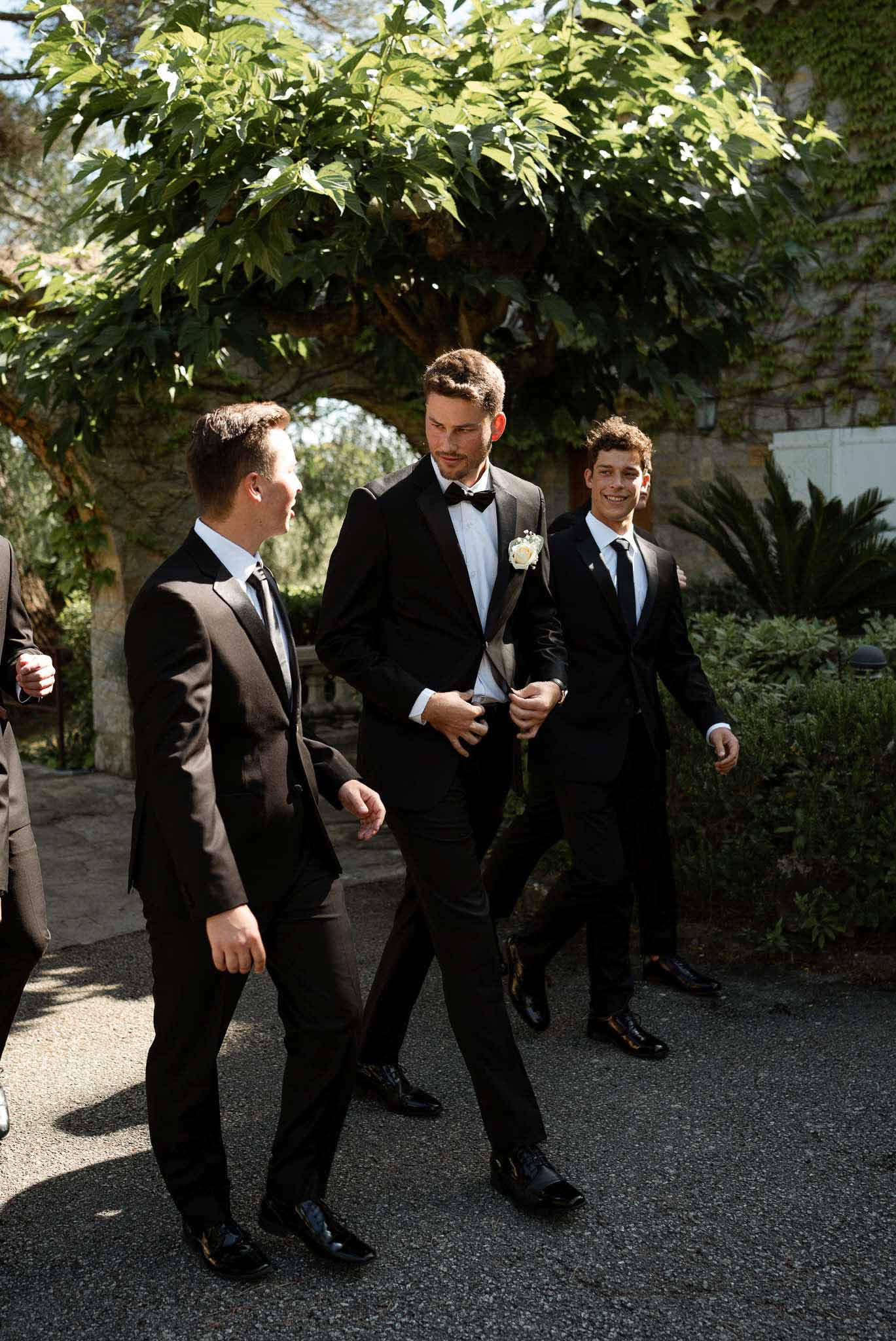 Groom in black tuxedo with white rose boutonniere walking gravel path with three groomsmen in black suits
