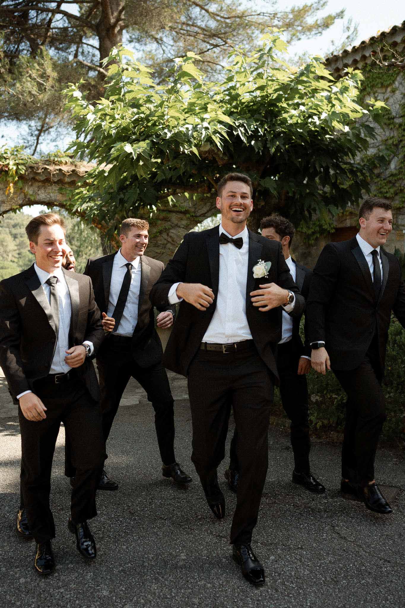Groom and five groomsmen in black suits laugh walking along paved path at vine-covered stone venue