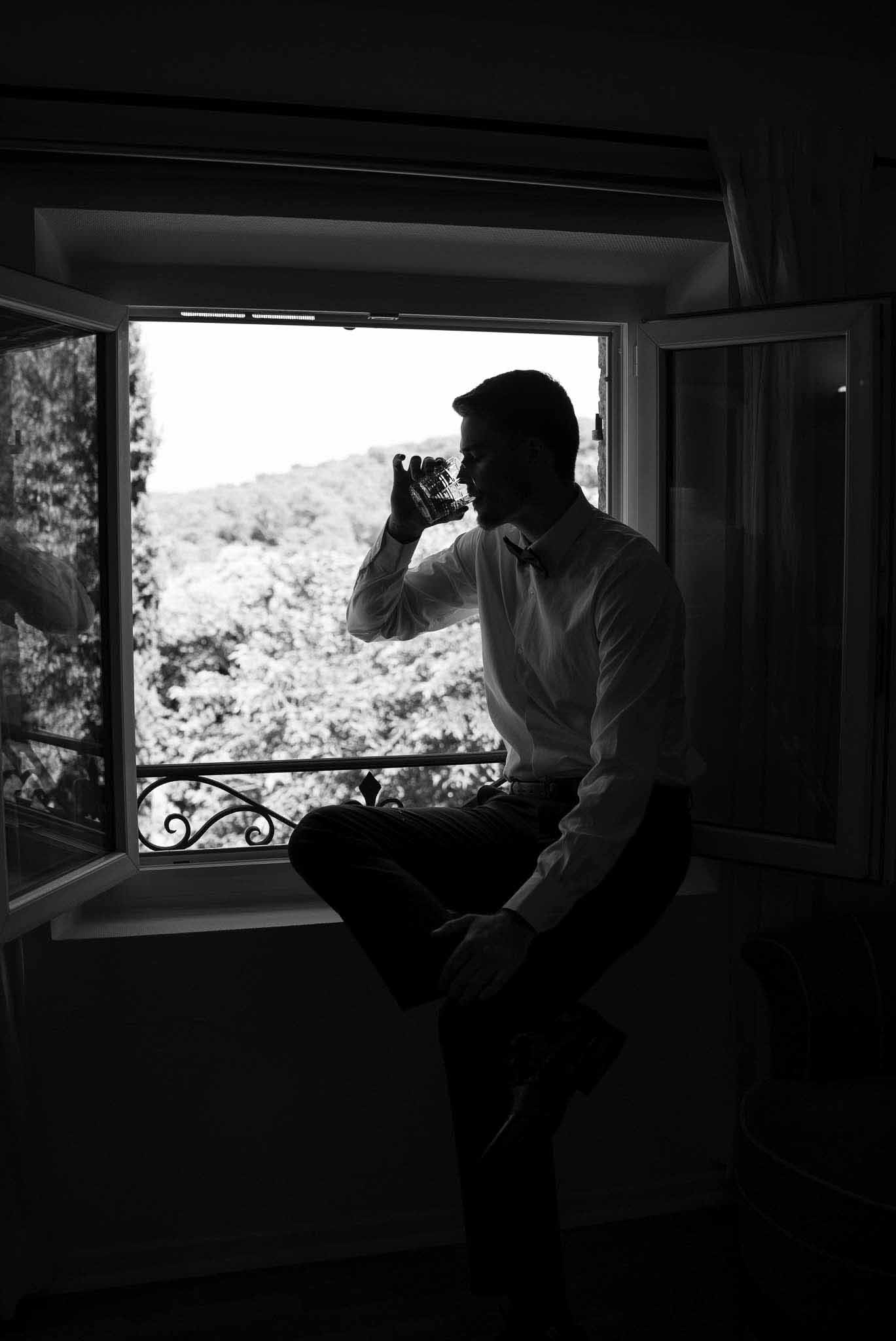 Black and white groom in suspenders and bow tie drinking from crystal glass on windowsill with wrought iron balcony