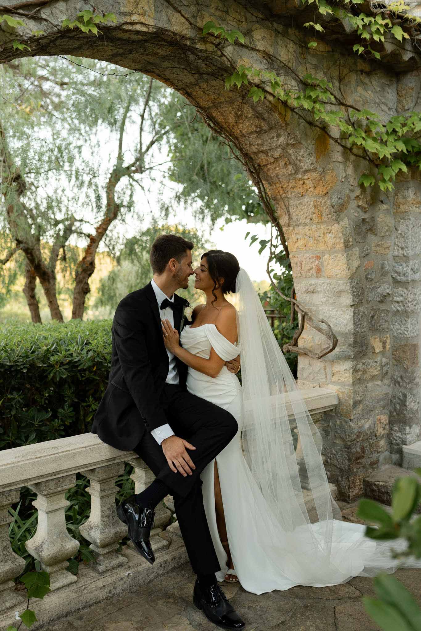 Groom on stone balustrade with bride in off-shoulder gown and cathedral veil at vine-covered terrace