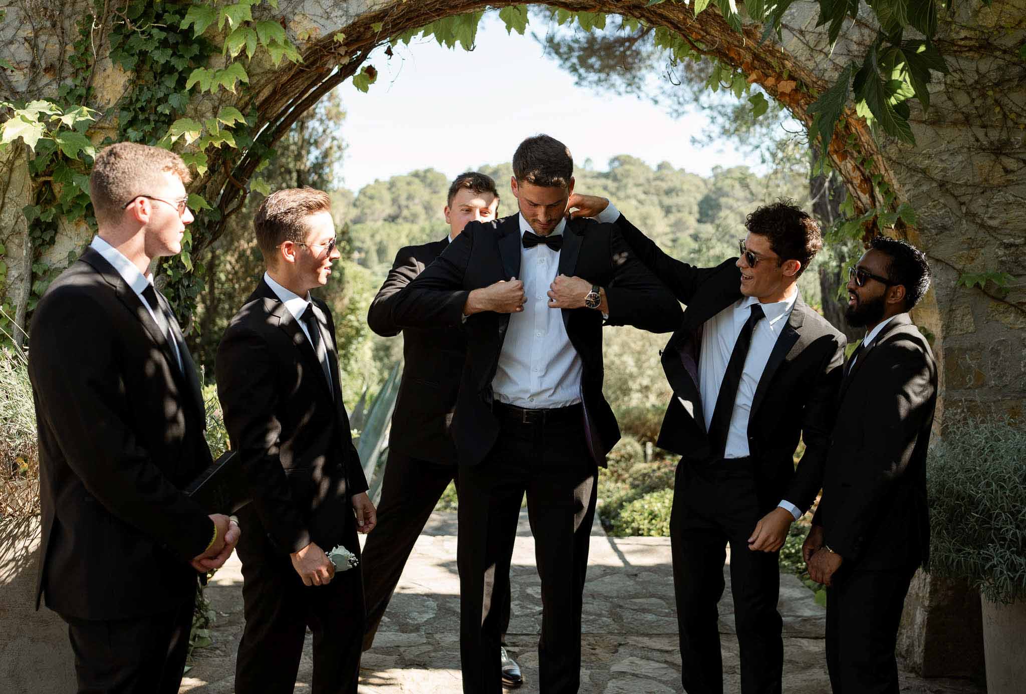 Groom being helped into black tuxedo jacket by groomsmen beneath a vine-covered stone archway outdoors