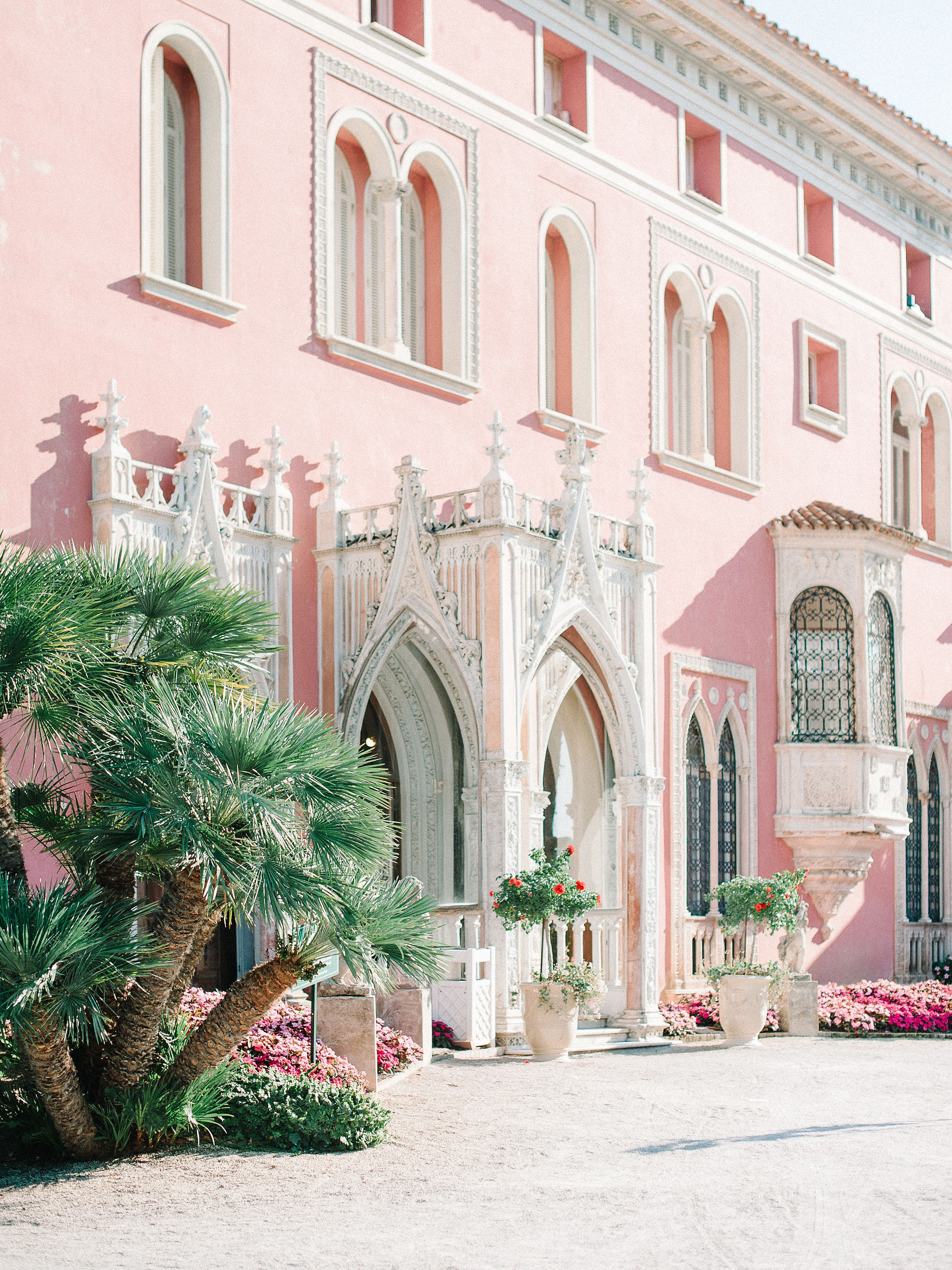 Salmon-pink villa facade with white Gothic arched doorways, stone urns with topiary, and palm tree on gravel forecourt
