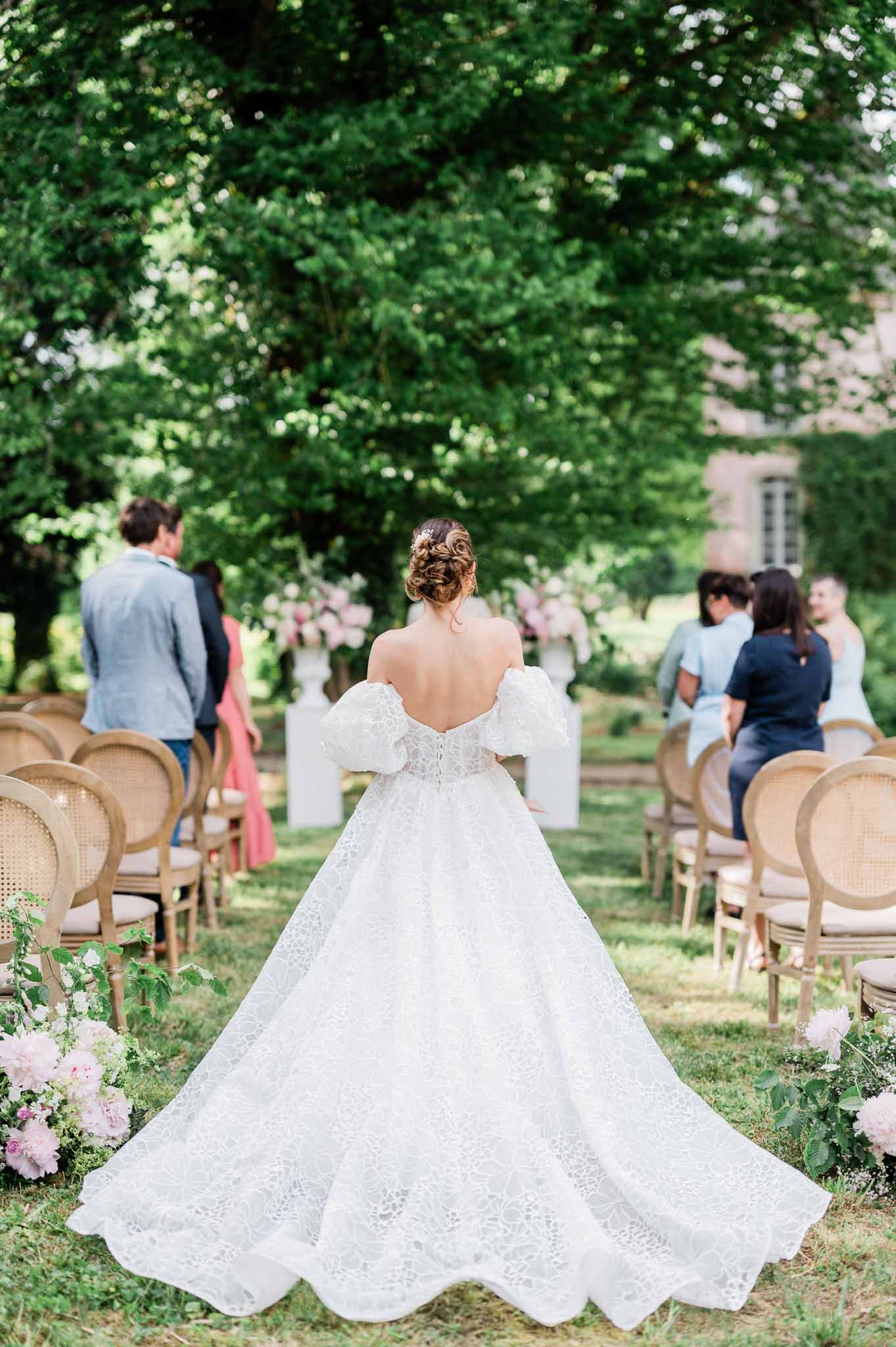 Bride walking down garden aisle during outdoor wedding ceremony with guests seated in wooden chairs