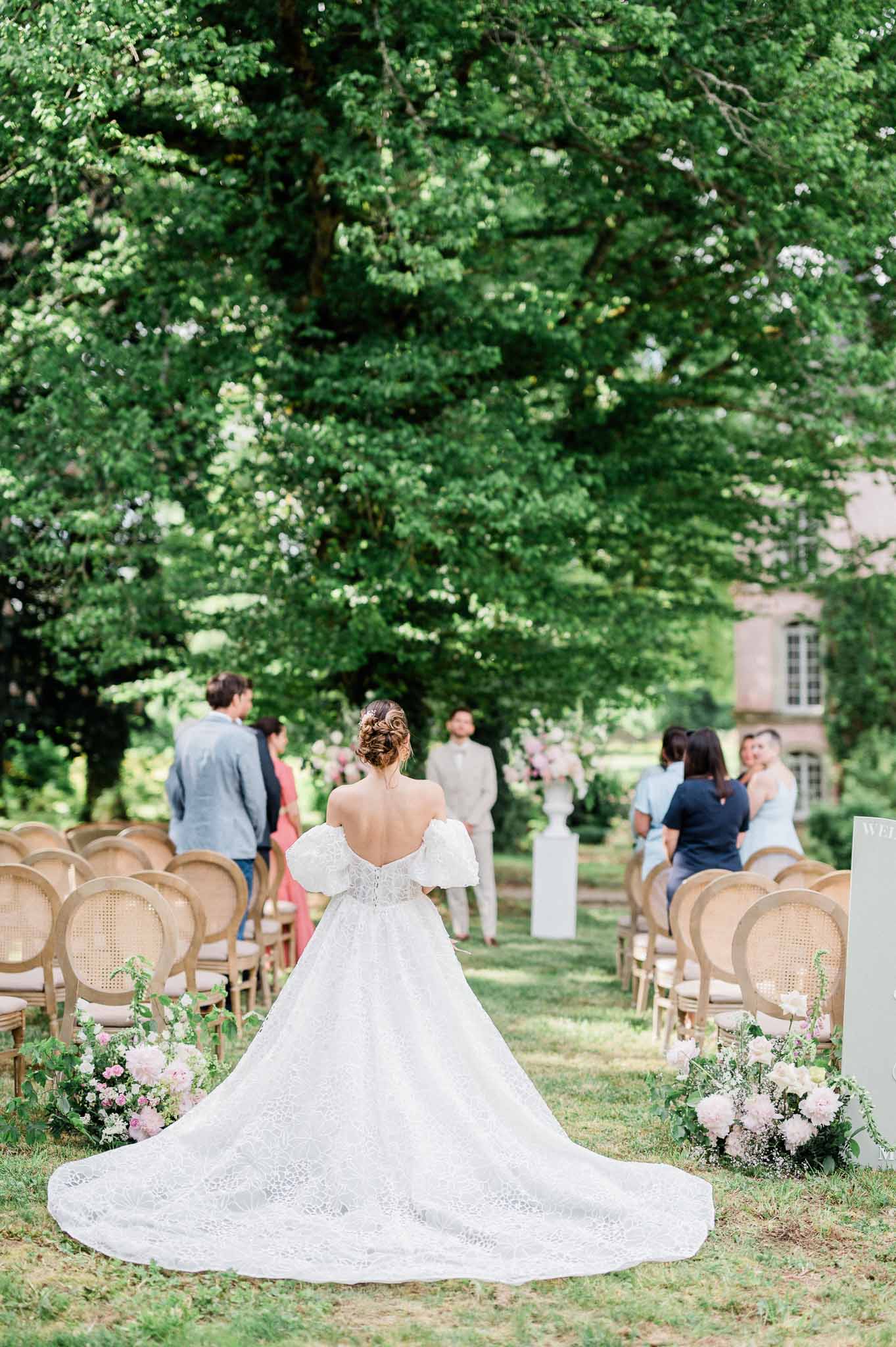 Bride walking down garden ceremony aisle at outdoor wedding venue with stone building backdrop