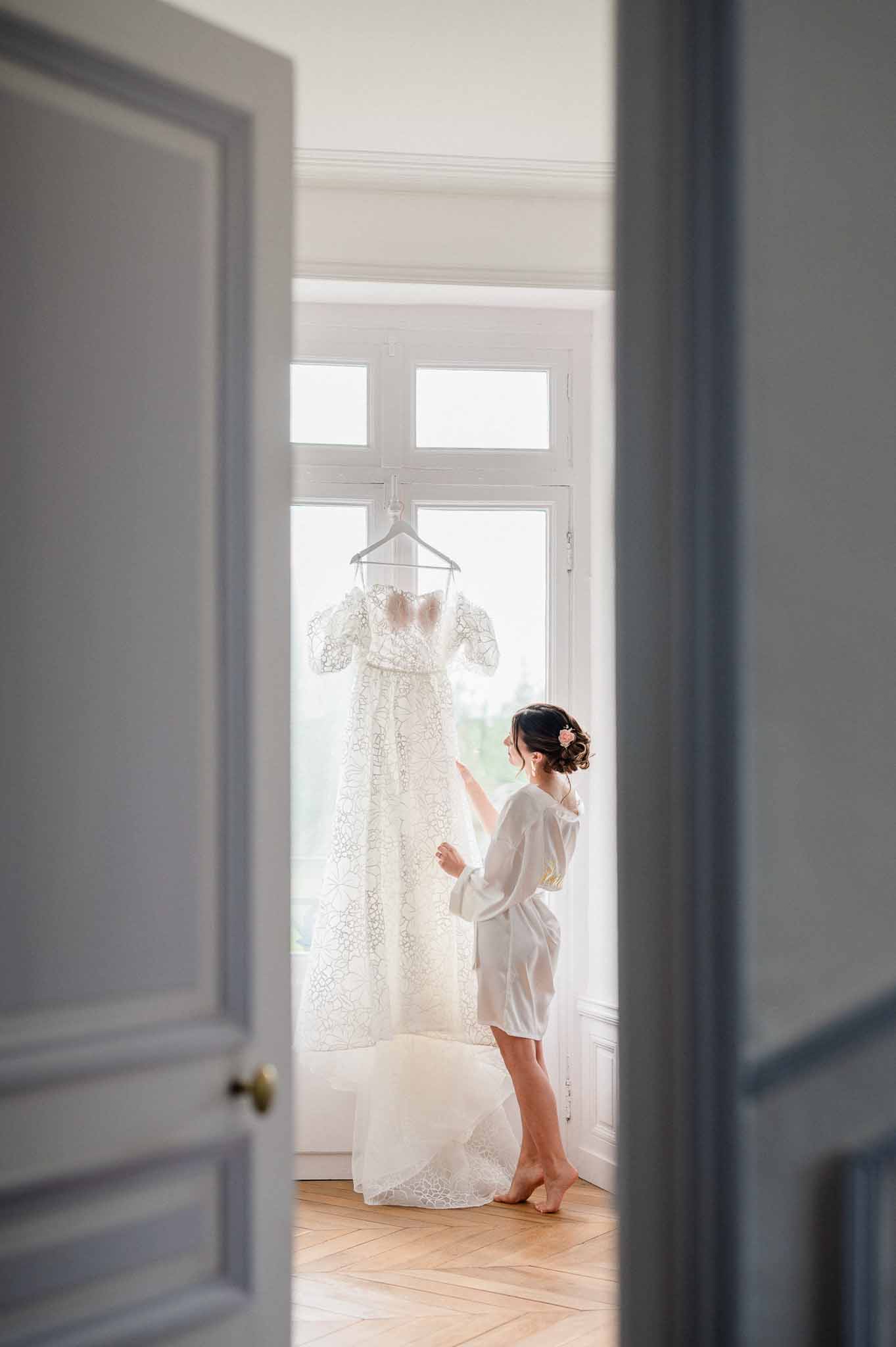 Bride in silk robe admiring hanging wedding dress in bright minimalist room with tall windows
