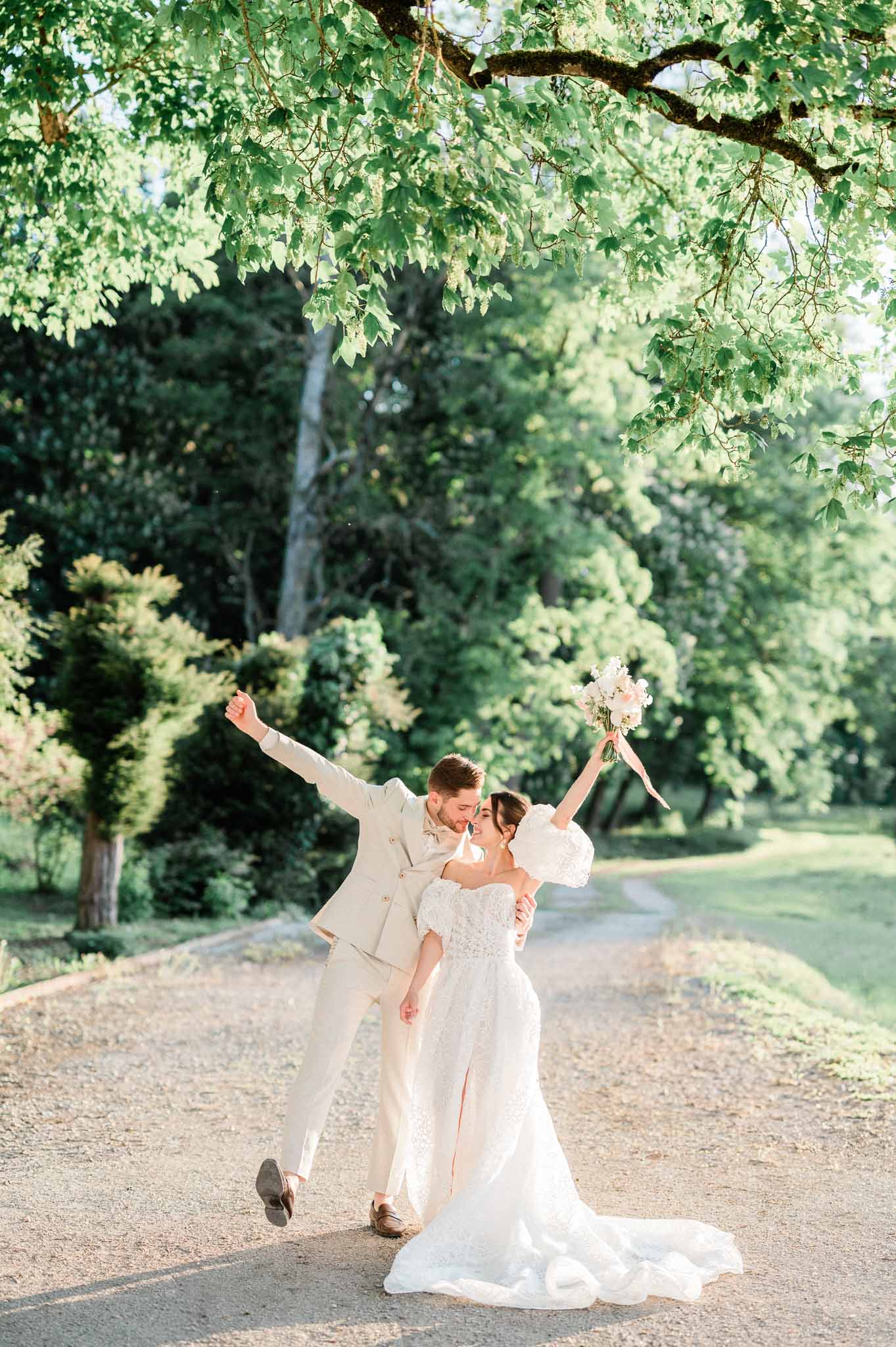 Groom lifts bride in celebratory pose on tree-lined pathway at estate garden wedding