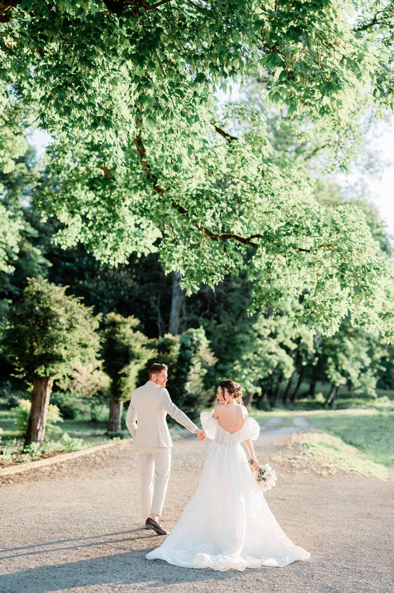 Bride and groom walking hand-in-hand down tree-lined pathway at garden wedding venue