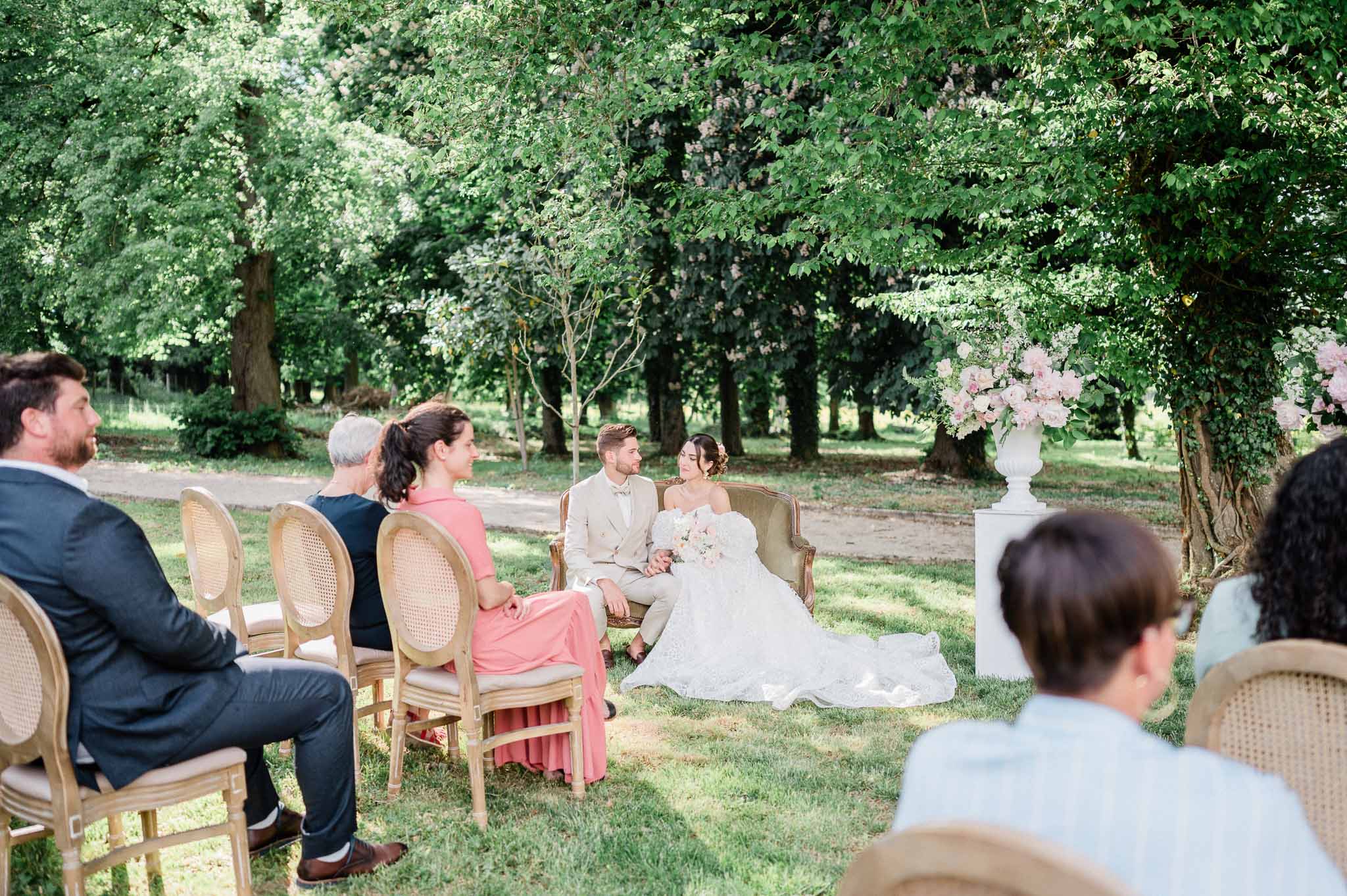 Outdoor garden wedding ceremony with couple and guests seated among mature trees and floral arrangements