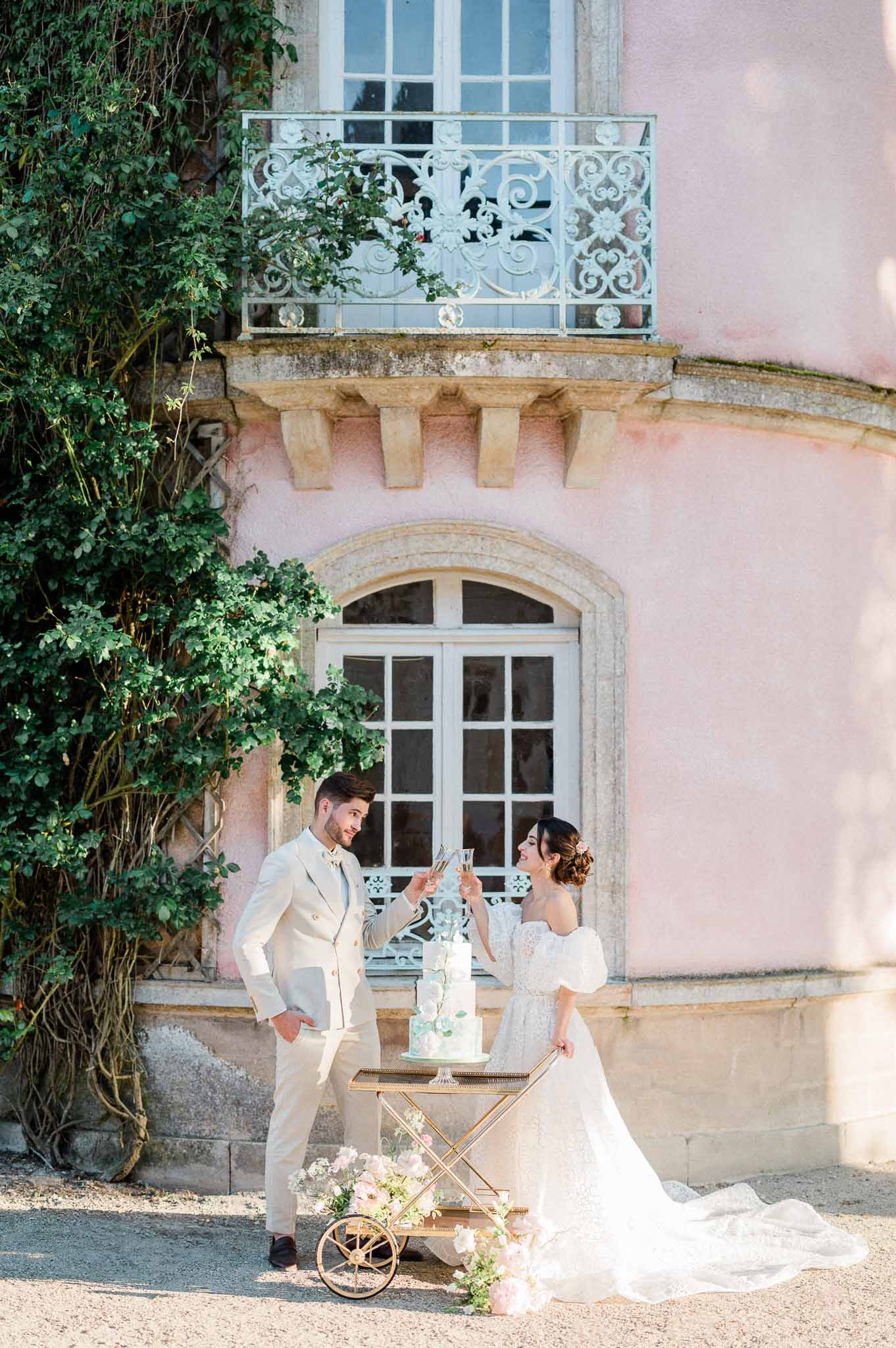 Bride and groom cutting wedding cake in front of pink château with ornate balconies