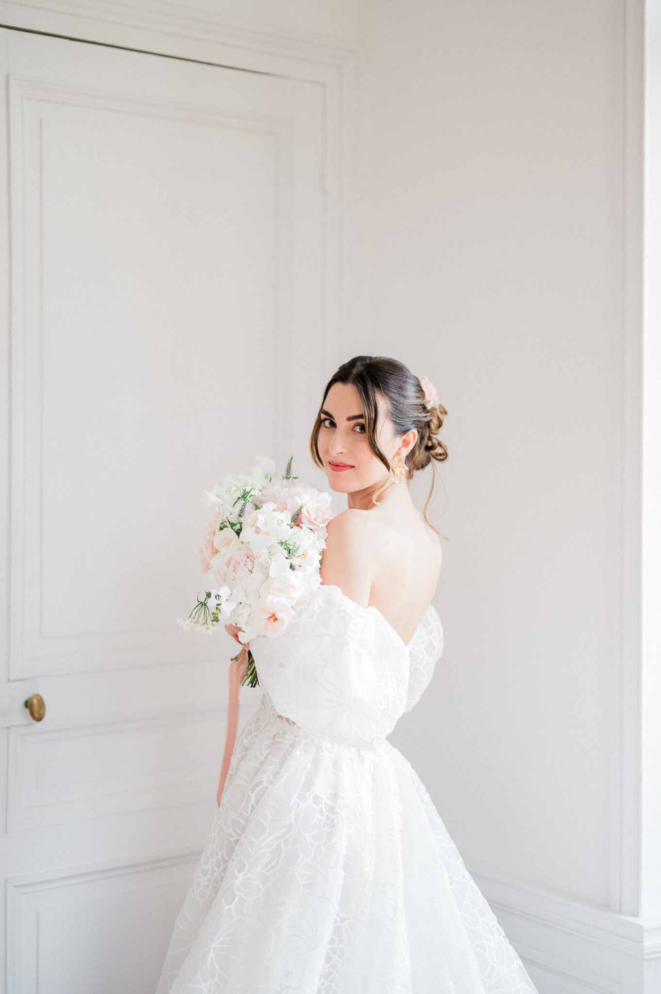 Bride in ivory lace wedding dress holding blush bouquet in minimalist white interior space