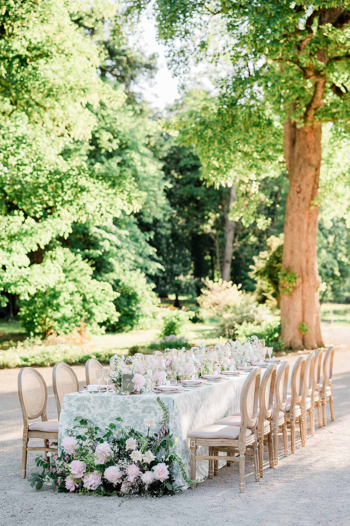 Outdoor reception table setup with floral centerpieces in tree-lined garden setting
