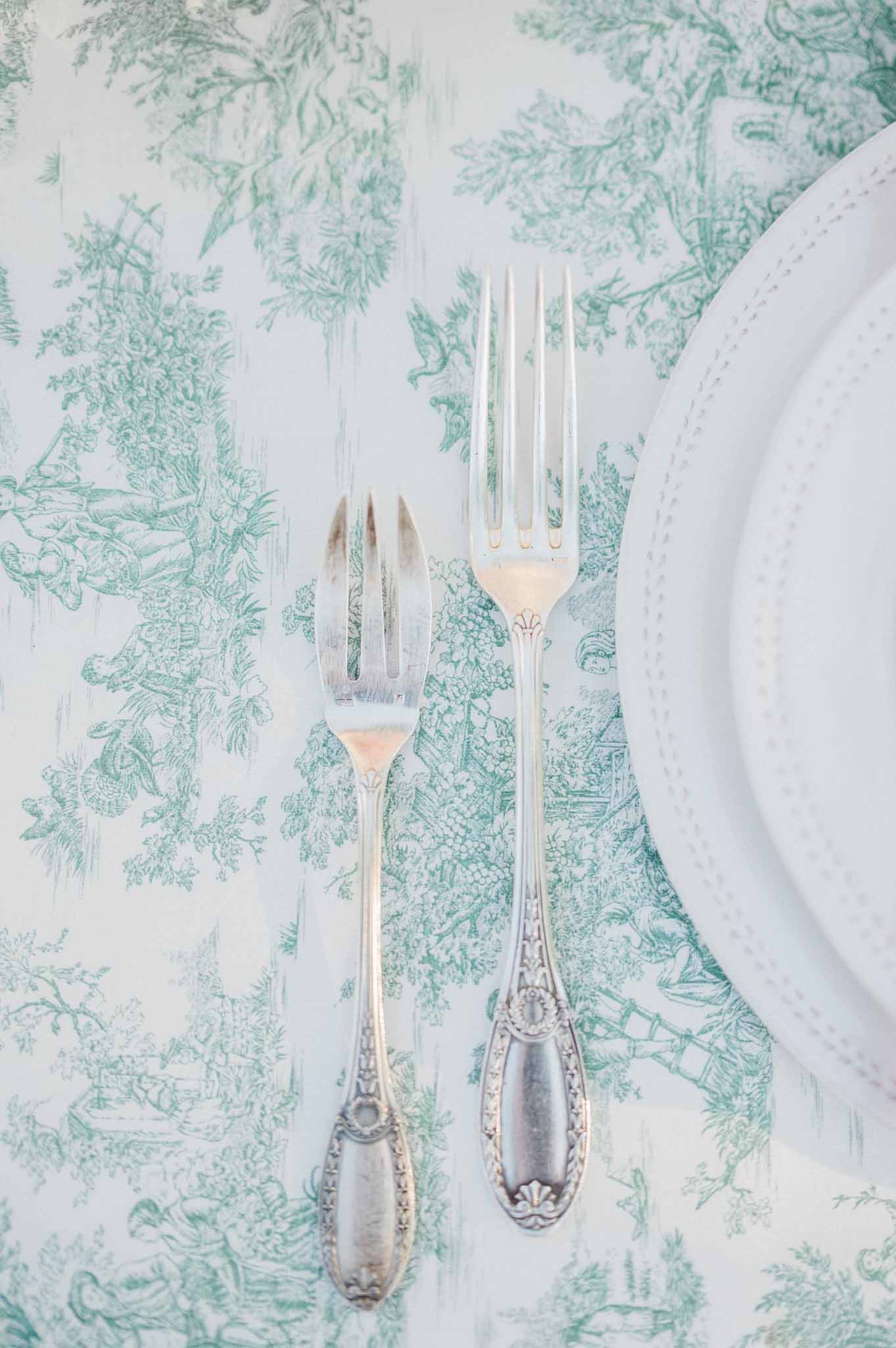 Ornate silver forks on green toile tablecloth with white beaded rim plate at elegant wedding reception table