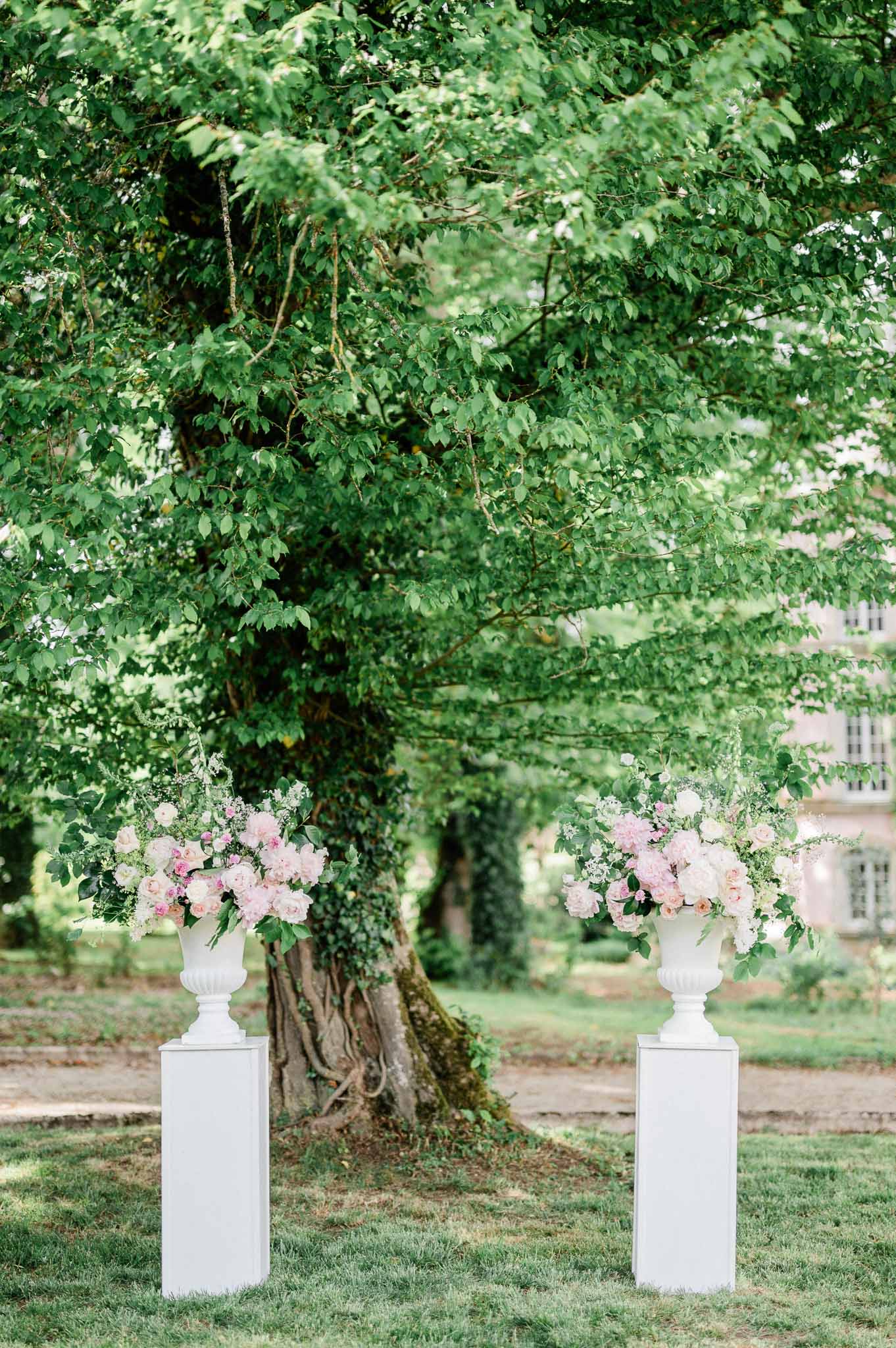 White neoclassical urns with blush pink roses flanking tree in garden courtyard ceremony setting
