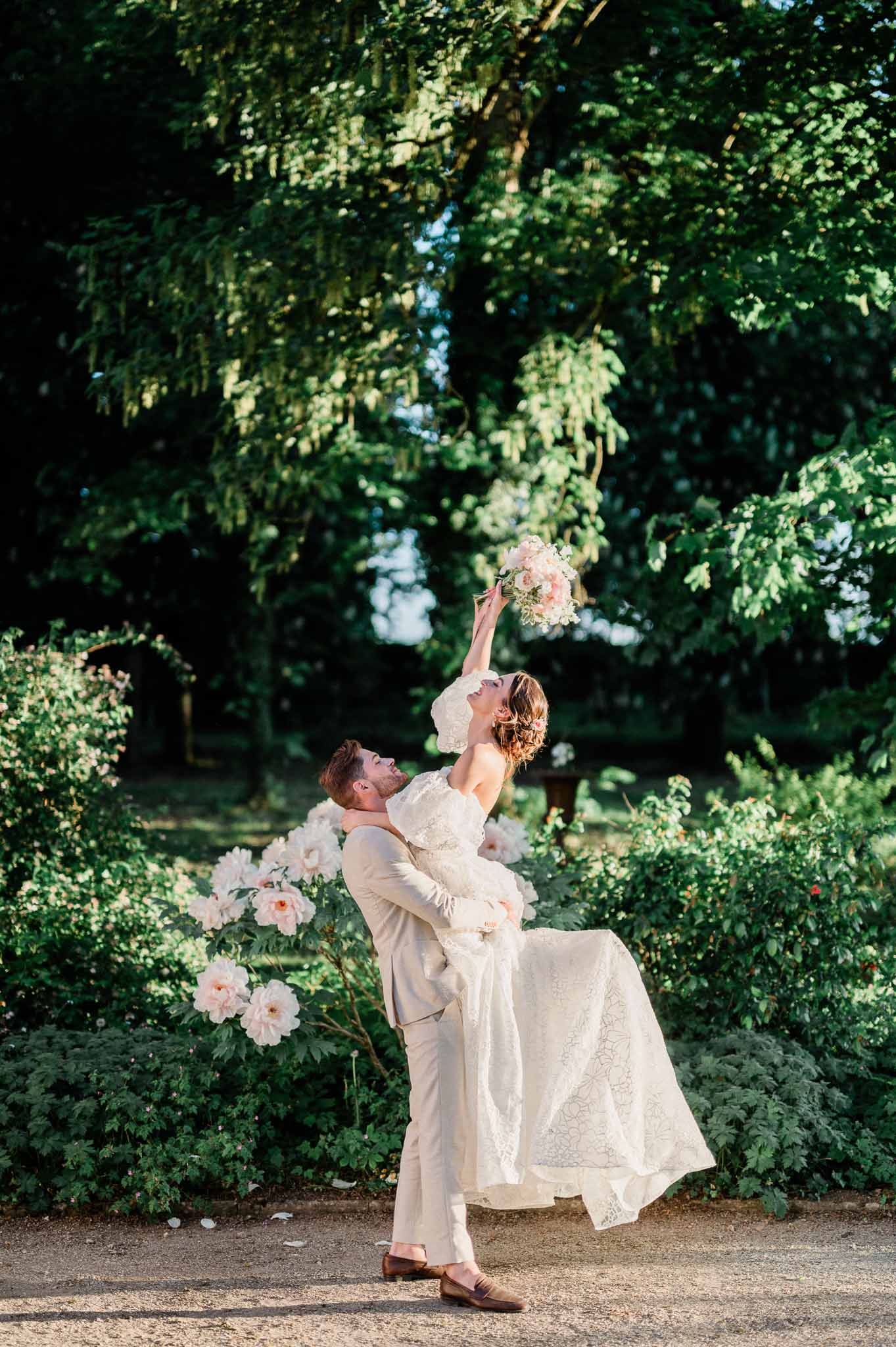Groom lifting bride in playful portrait moment in lush garden setting with dappled sunlight