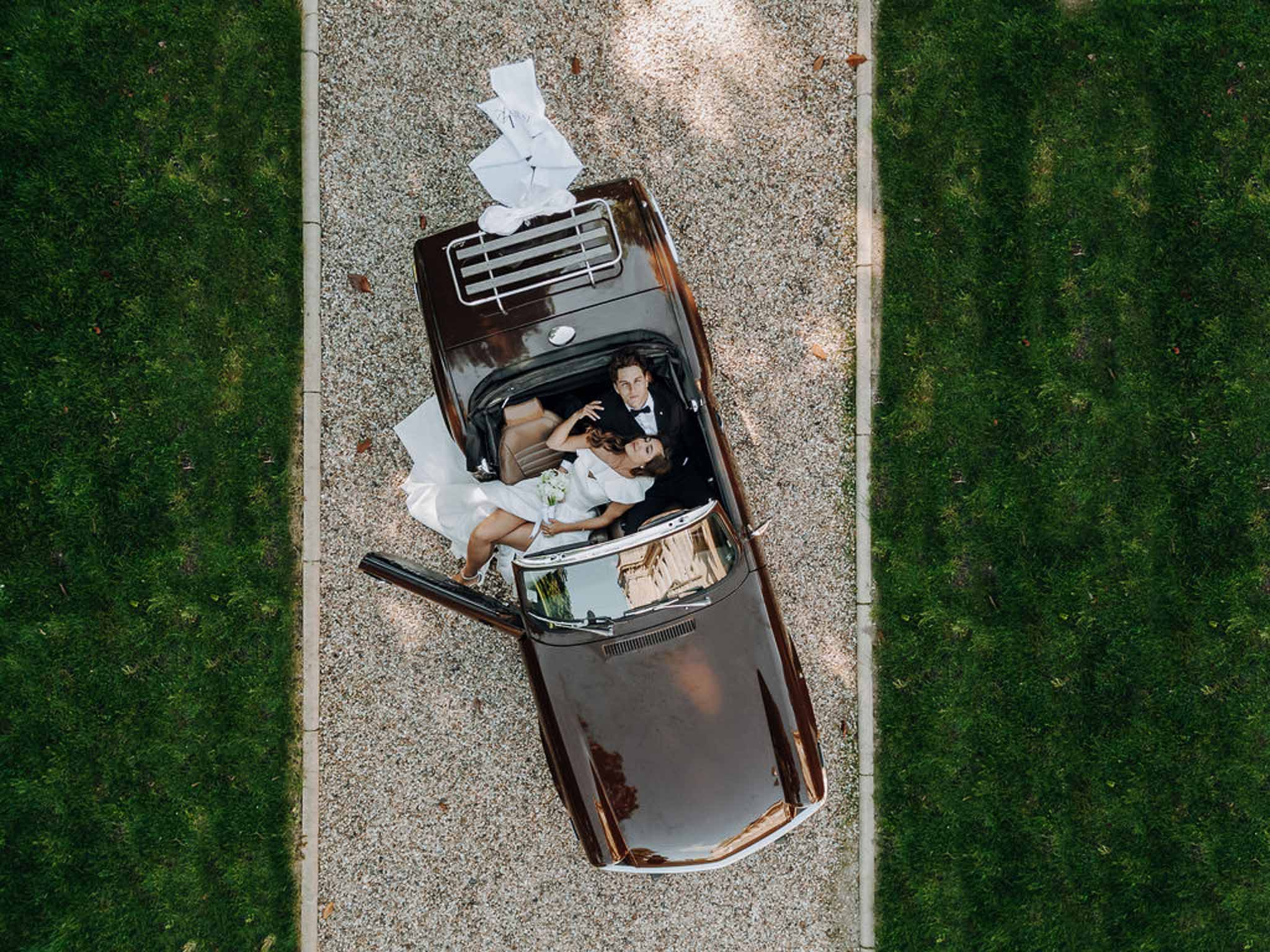 Top-down aerial of couple in vintage convertible on gravel, bride in white gown with bouquet and white ribbon
