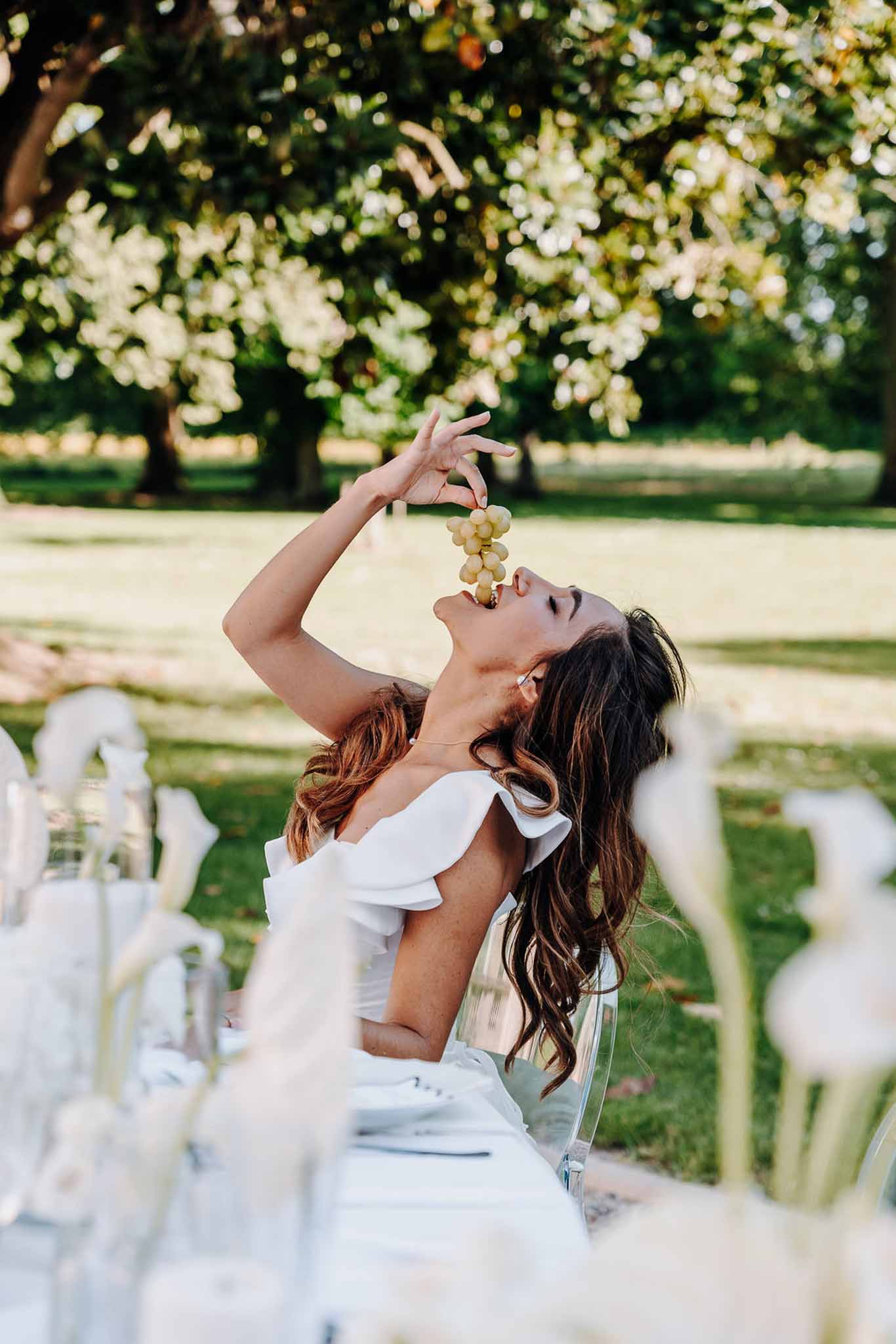 Bride in off-shoulder ruffled gown playfully eating grapes at outdoor reception table