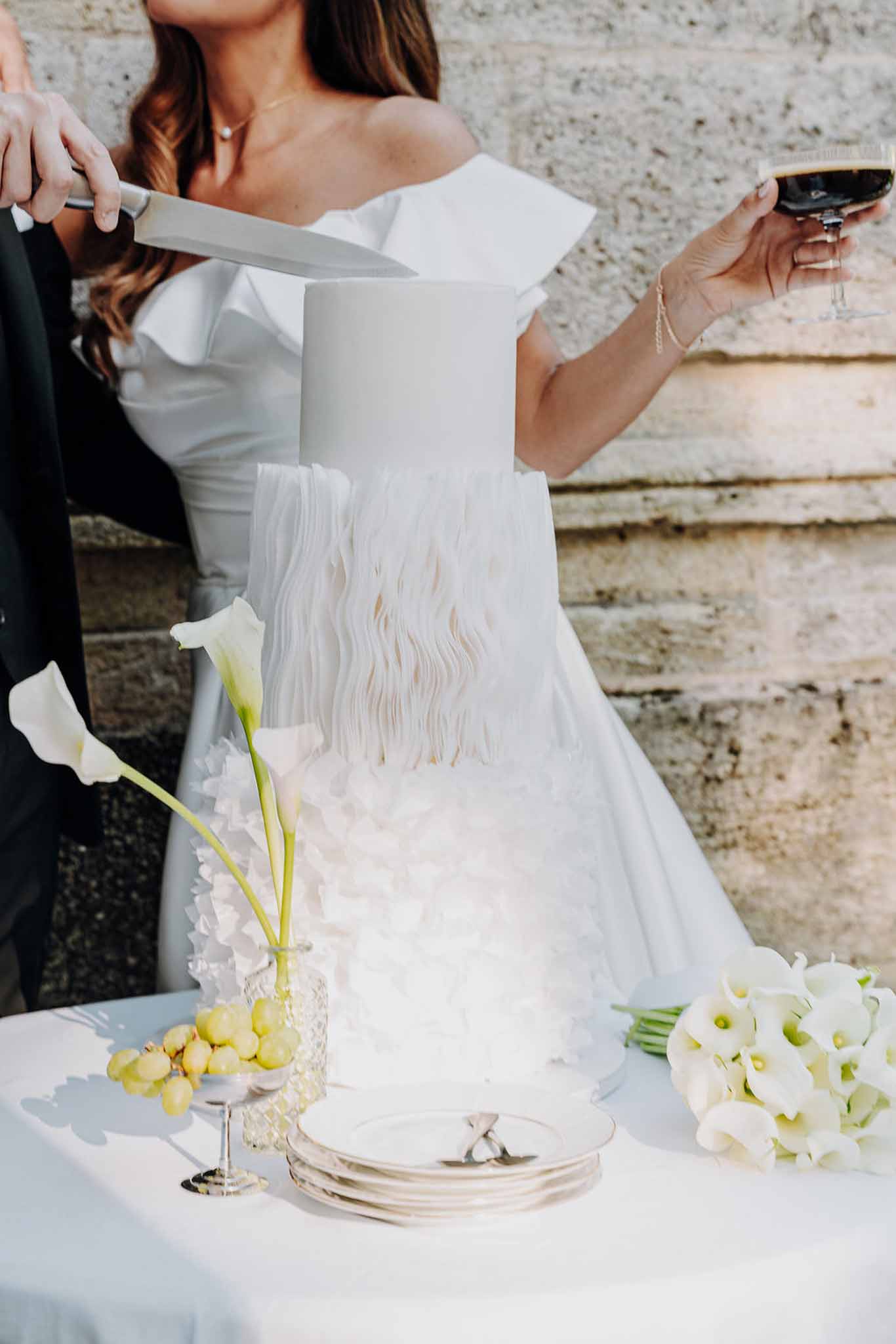 Bride and groom cutting three-tier white ruffled wedding cake outdoors with calla lilies on table