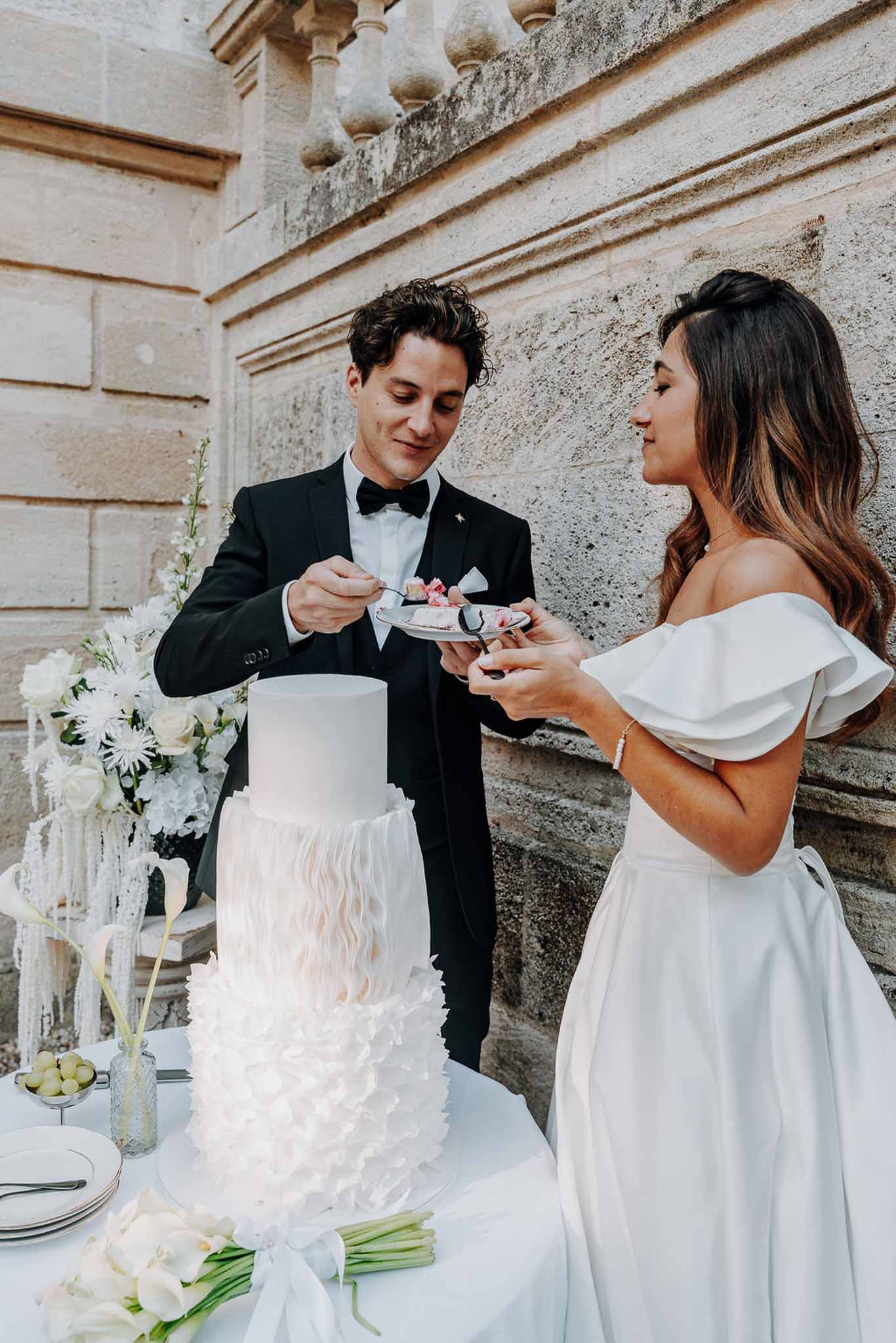 Bride and groom taste their three-tier white ruffled wedding cake outside a stone chateau facade