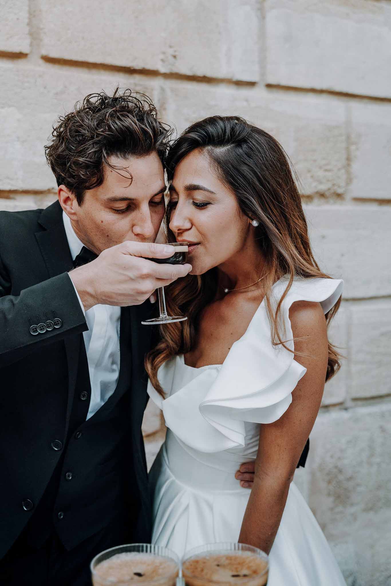 Couple sharing wine glass with heads pressed together against stone wall in ruffle-shoulder gown and tux