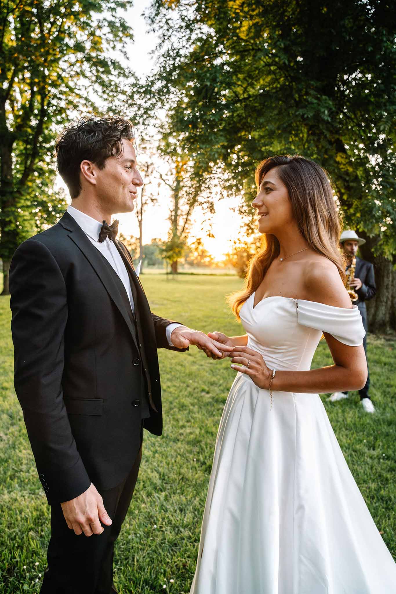 Couple holding hands at golden hour with saxophonist in soft-focus background among trees