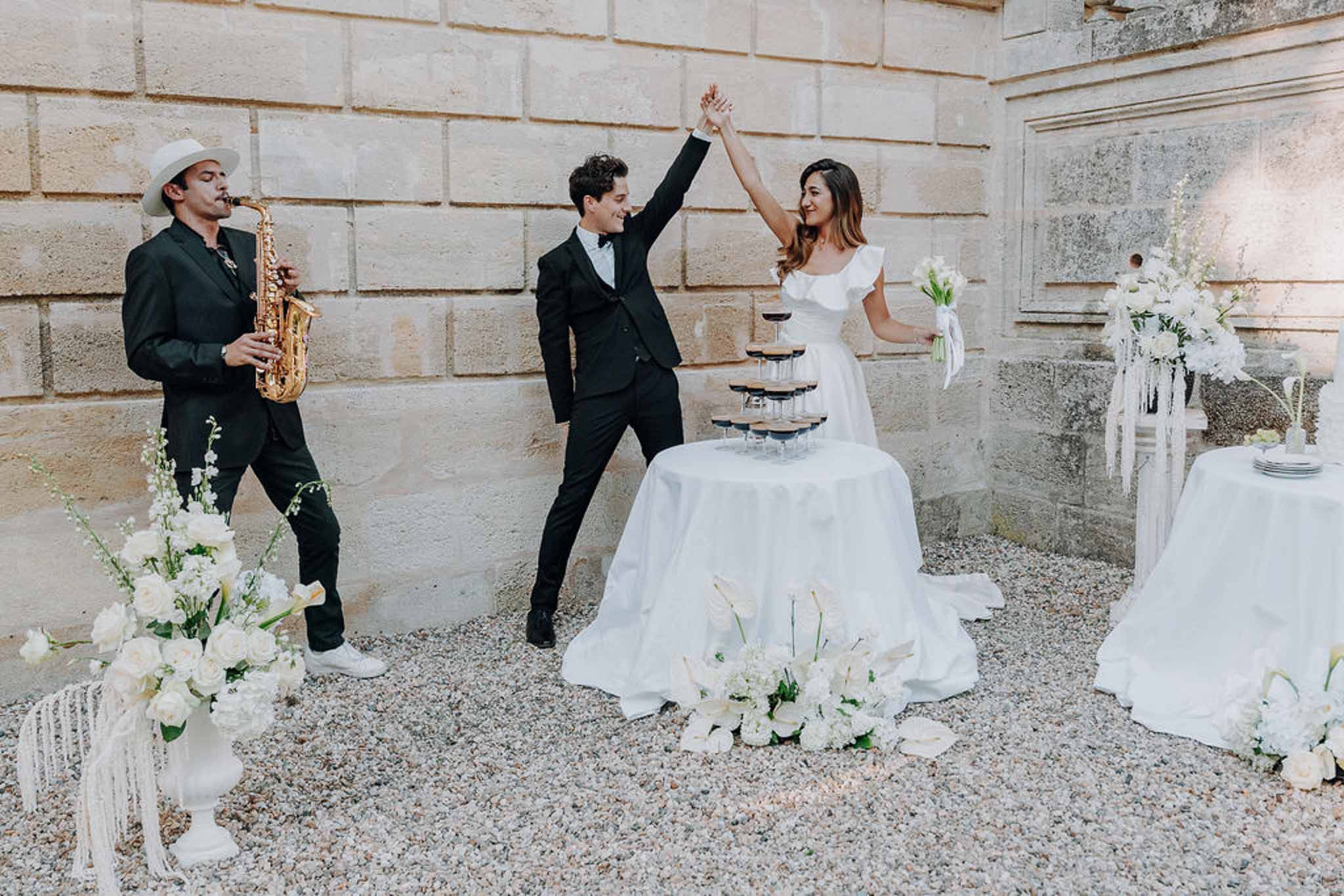 Bride and groom celebrating with saxophonist beside champagne tower and all-white floral arrangements