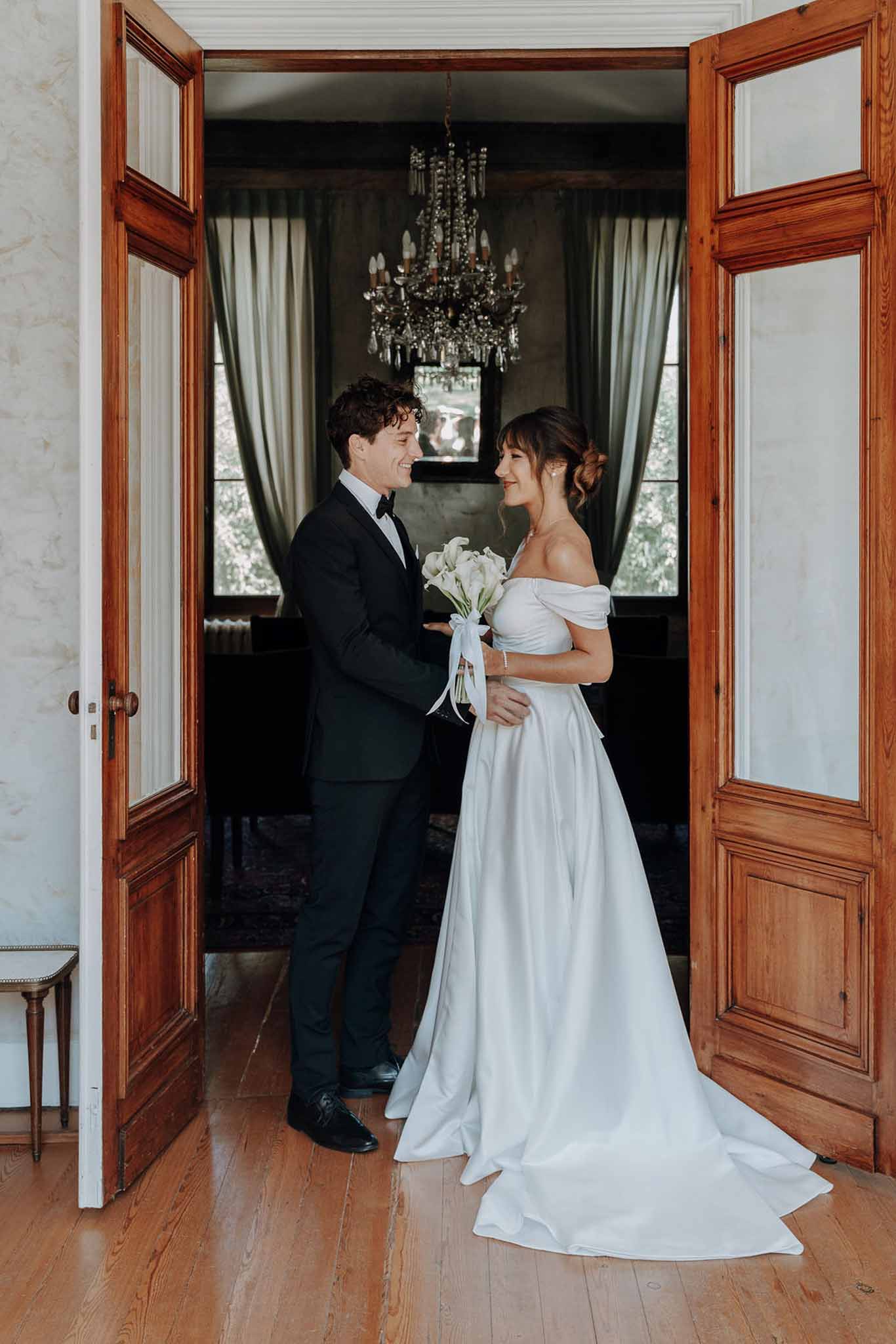 Couple in grand double doorway with bride holding white calla lily bouquet and chandelier beyond
